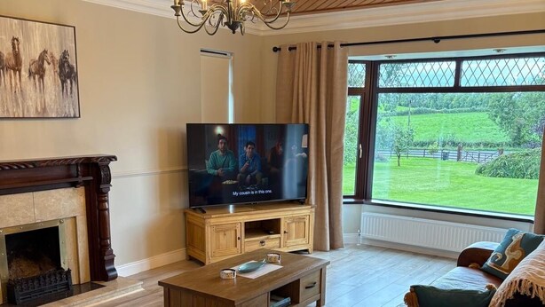 Ein Wohnzimmer mit Kamin, Fernseher und einem großen Fenster mit Blick auf eine grüne Landschaft.