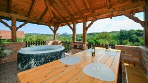 A wooden pergola with a hot tub and a table set for dining, overlooking a lush landscape.