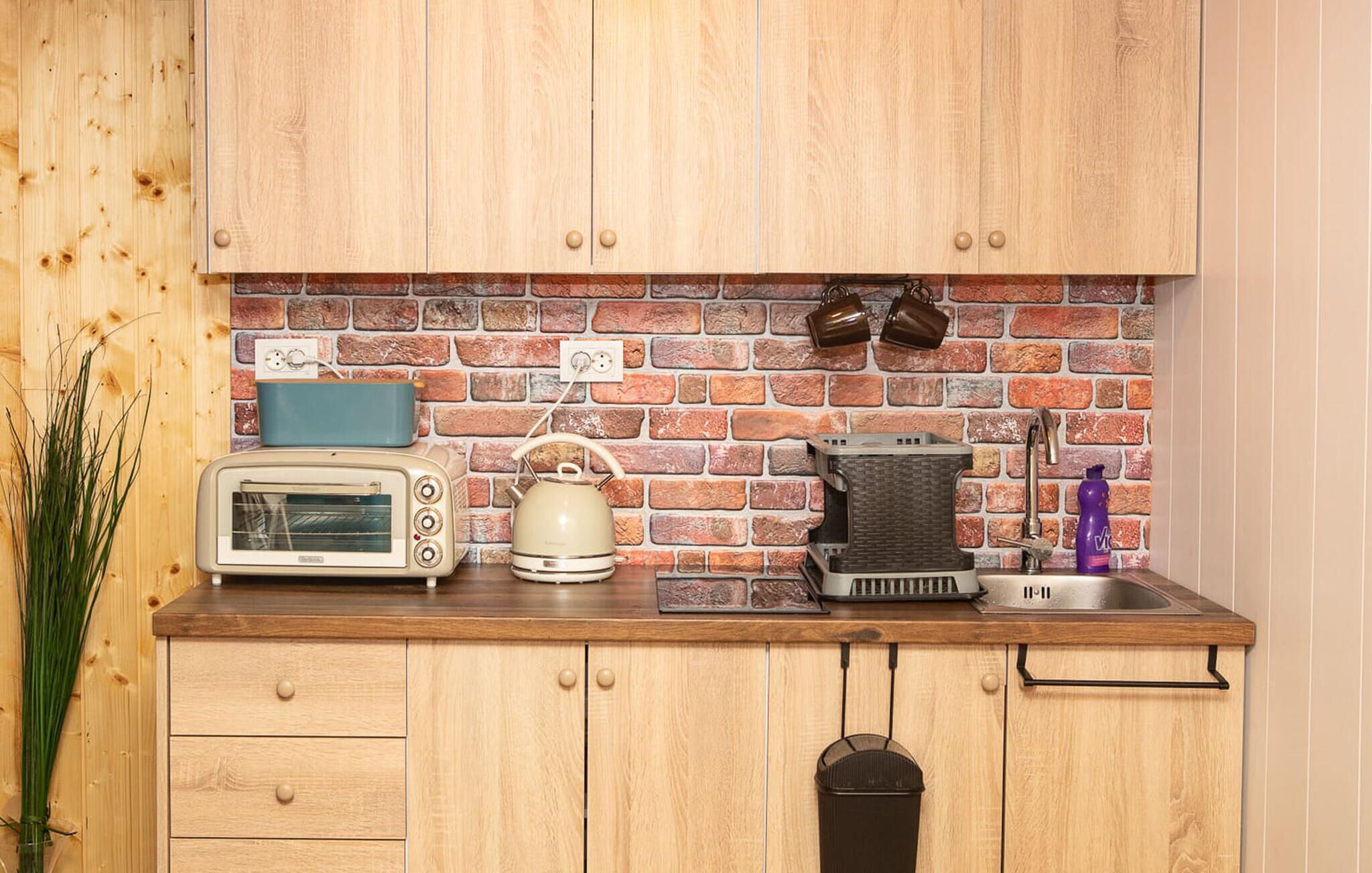 A kitchen with wooden cabinets, a brick backsplash, a toaster, a kettle, a sink, and a vase with green plants.