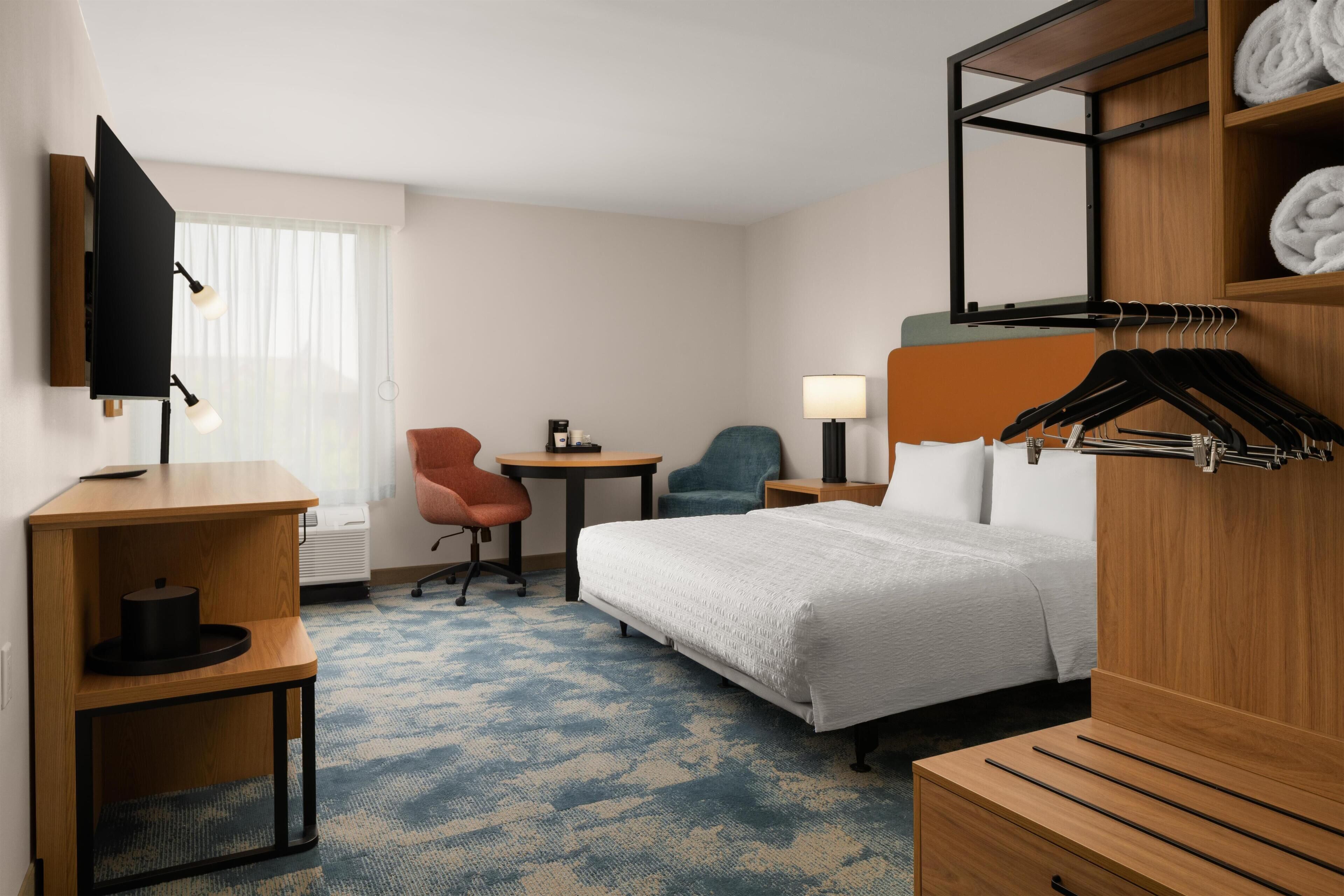 A hotel room with a bed, a desk, a chair, a TV, a lamp, and a wardrobe with hangers.