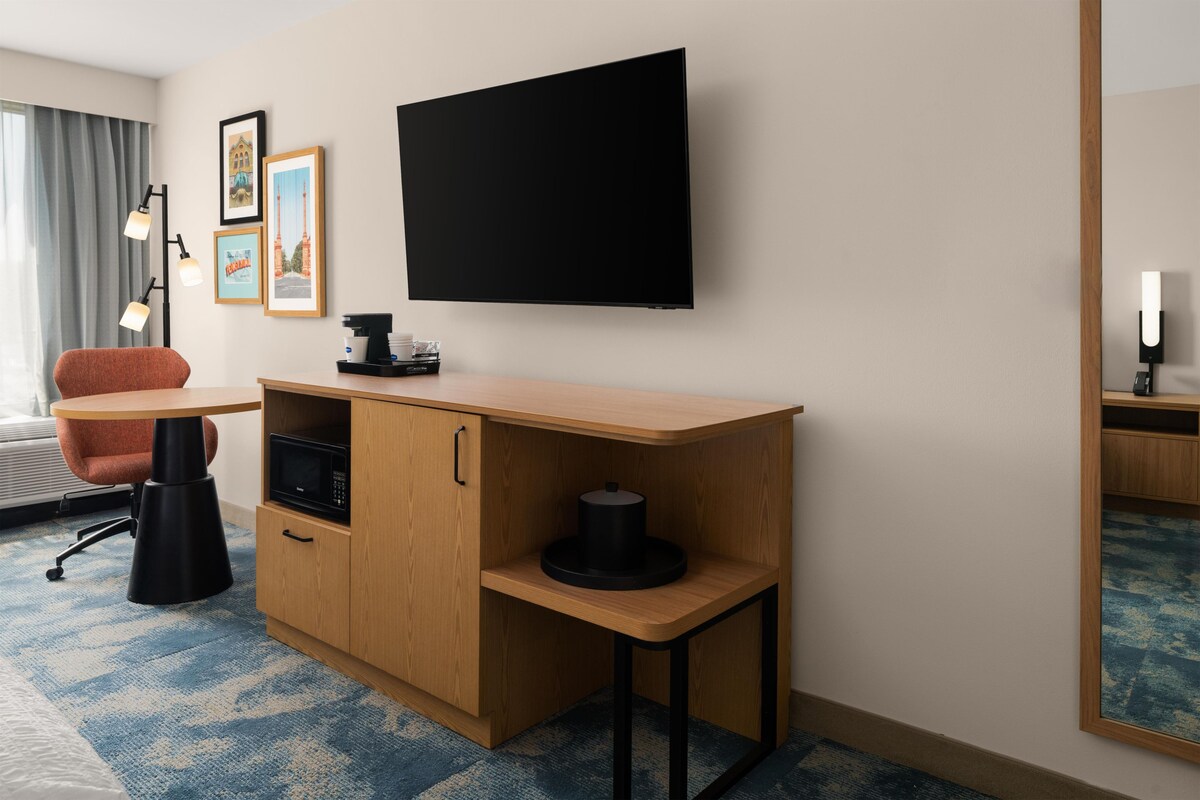A hotel room with a wooden desk, a flat-screen TV, a coffee machine, a microwave, a chair, and framed pictures on the wall.