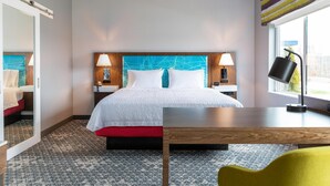 Ein Hotelzimmer mit Bett, Nachttischen, Schreibtisch, Stuhl und einem fensterbelaubten Fenster.