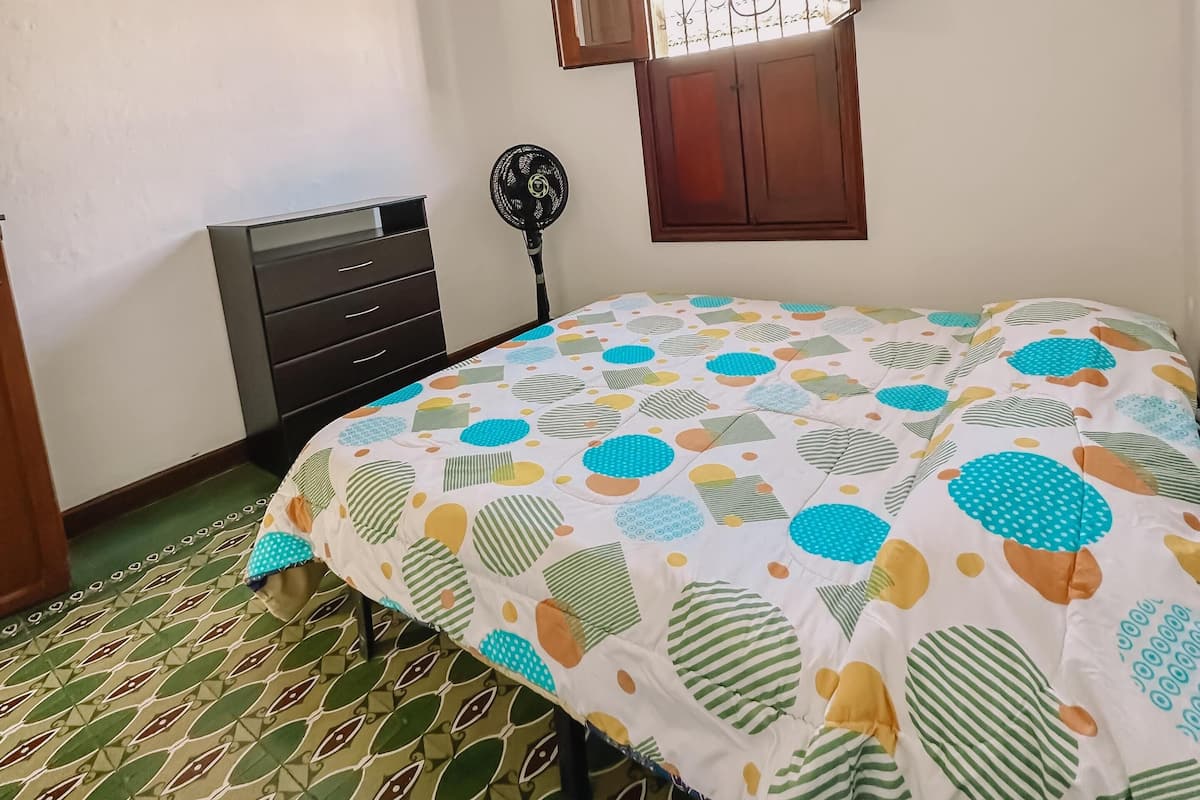 Une chambre à coucher comprenant un lit, une commode, un luminaire suspendu, une fenêtre avec des barreaux et un sol à motifs.