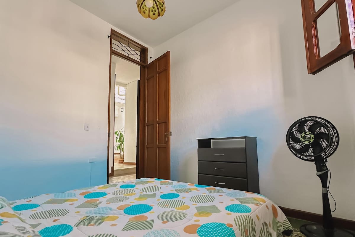 Une chambre à coucher comprenant un lit, une commode, un ventilateur sur pied et une porte donnant sur une autre pièce.