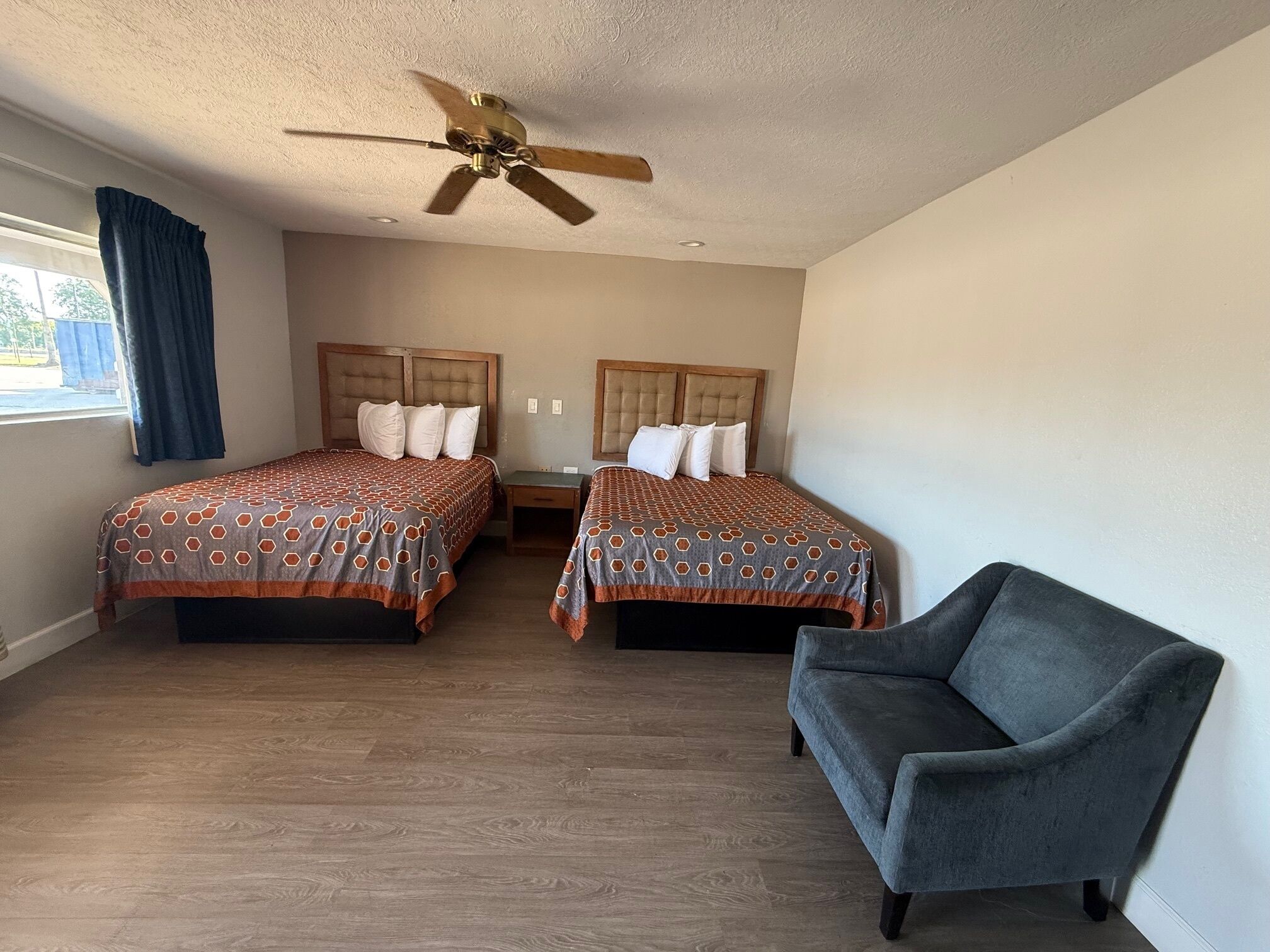 Una habitación de hotel con dos camas, un ventilador de techo, un sillón gris y una ventana con cortinas.