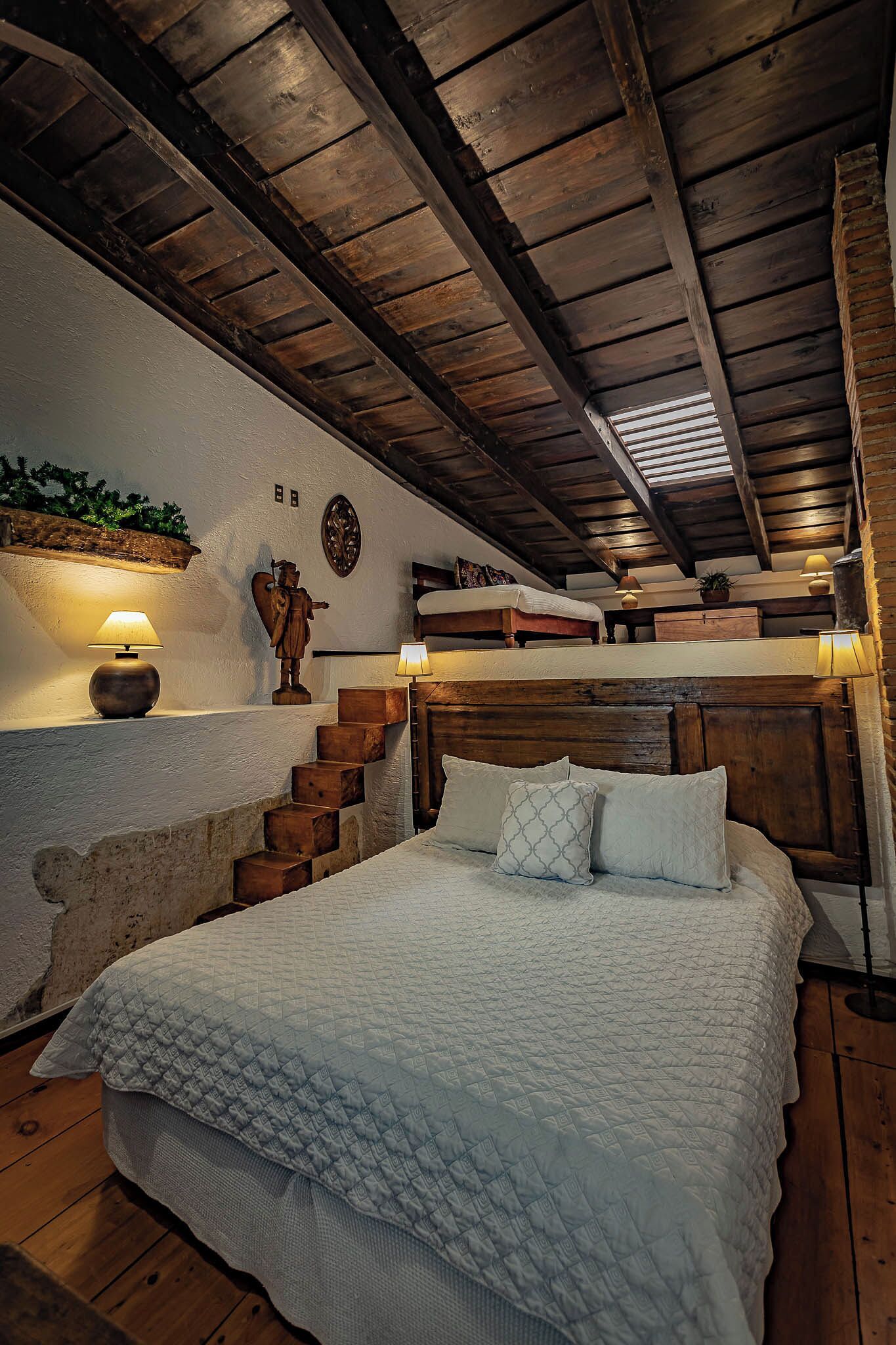 A bedroom with a wooden ceiling, a bed with white bedding, a lamp, a wooden chest, and a staircase.