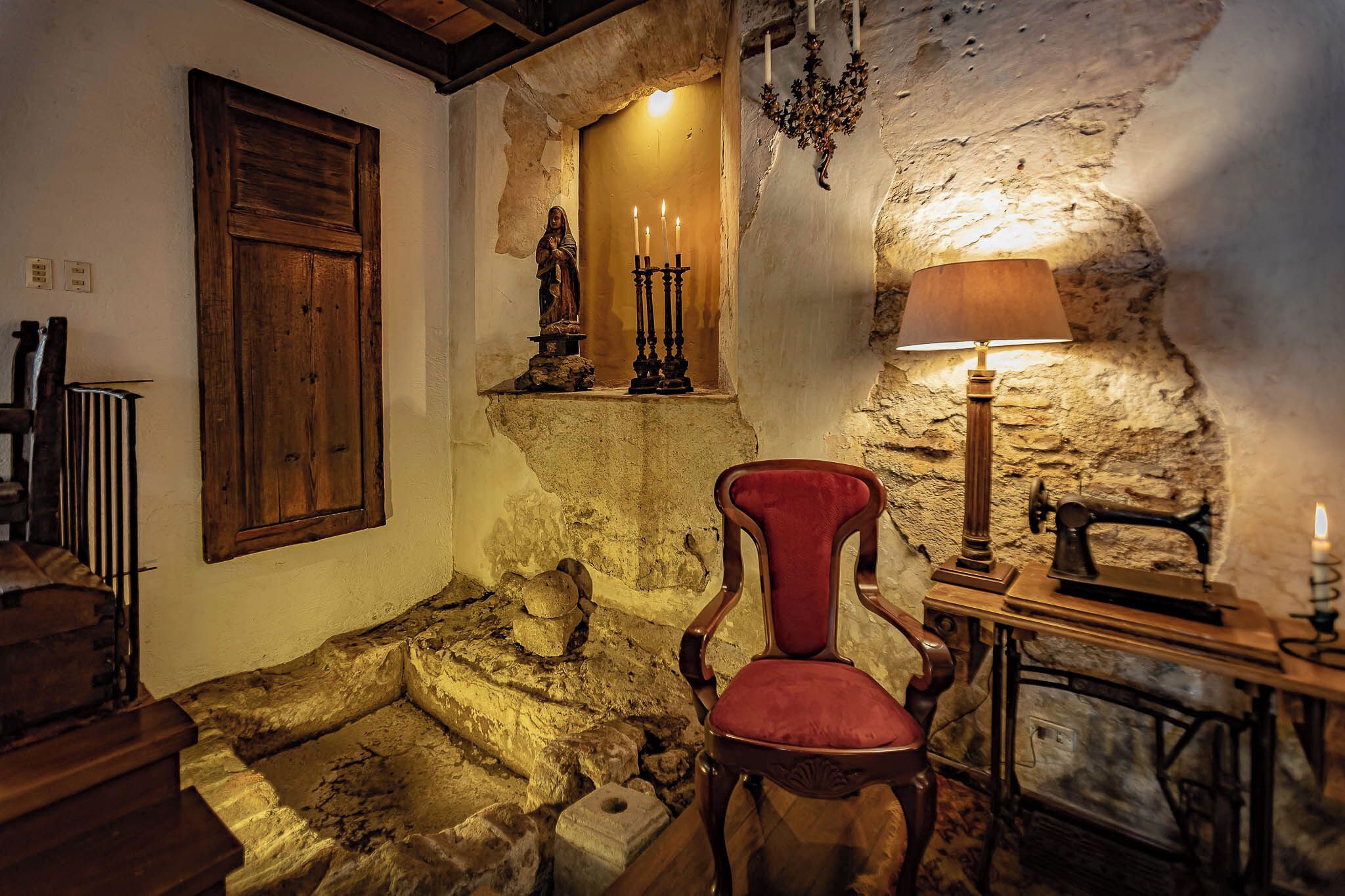 A rustic room with a wooden chair, a table with a lamp, a statue, and a stone wall.