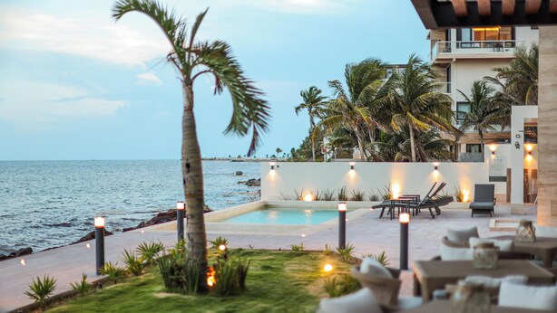 A poolside area with lounge chairs and tables, palm trees, and a view of the ocean. - Distrito Puerto - Ocean & Mangrove Views (Puerto Morelos)