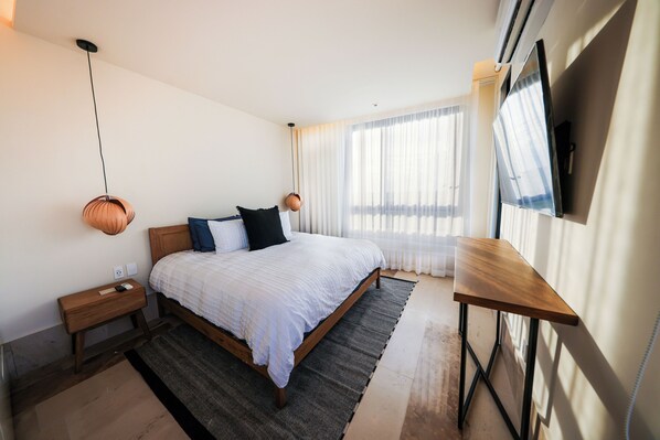 A modern bedroom with a wooden bed, a bedside table, a desk, and a large window with sheer curtains.