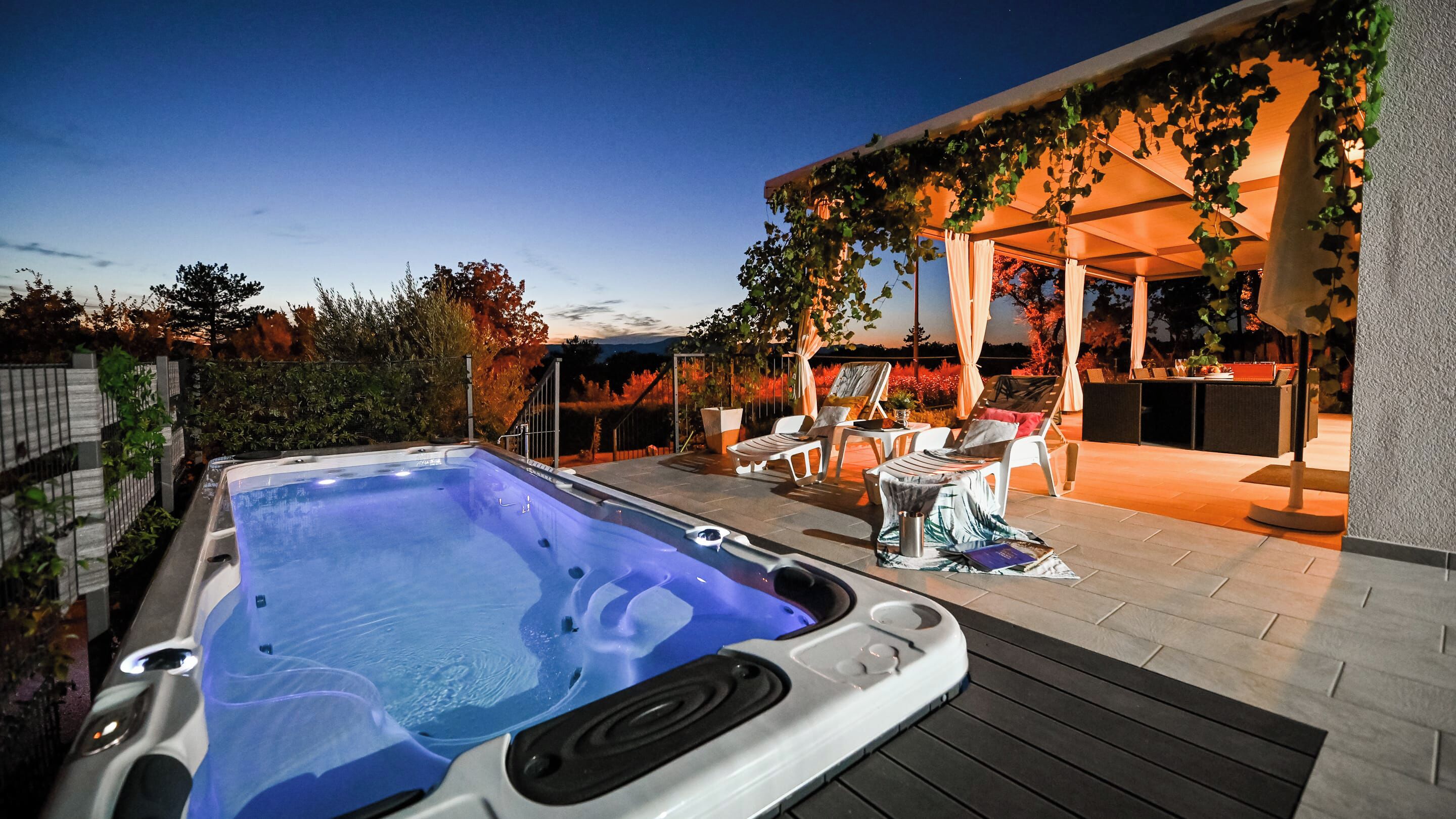 Een hottub met een houten terras, buitenmeubels en een pergola begroeid met wijnranken.