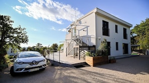 A modern two-story building with a car parked in front, a staircase leading to the upper floor, and a satellite dish on the roof.
