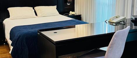 A hotel room with a bed, a desk, a chair, a nightstand, and a lamp.