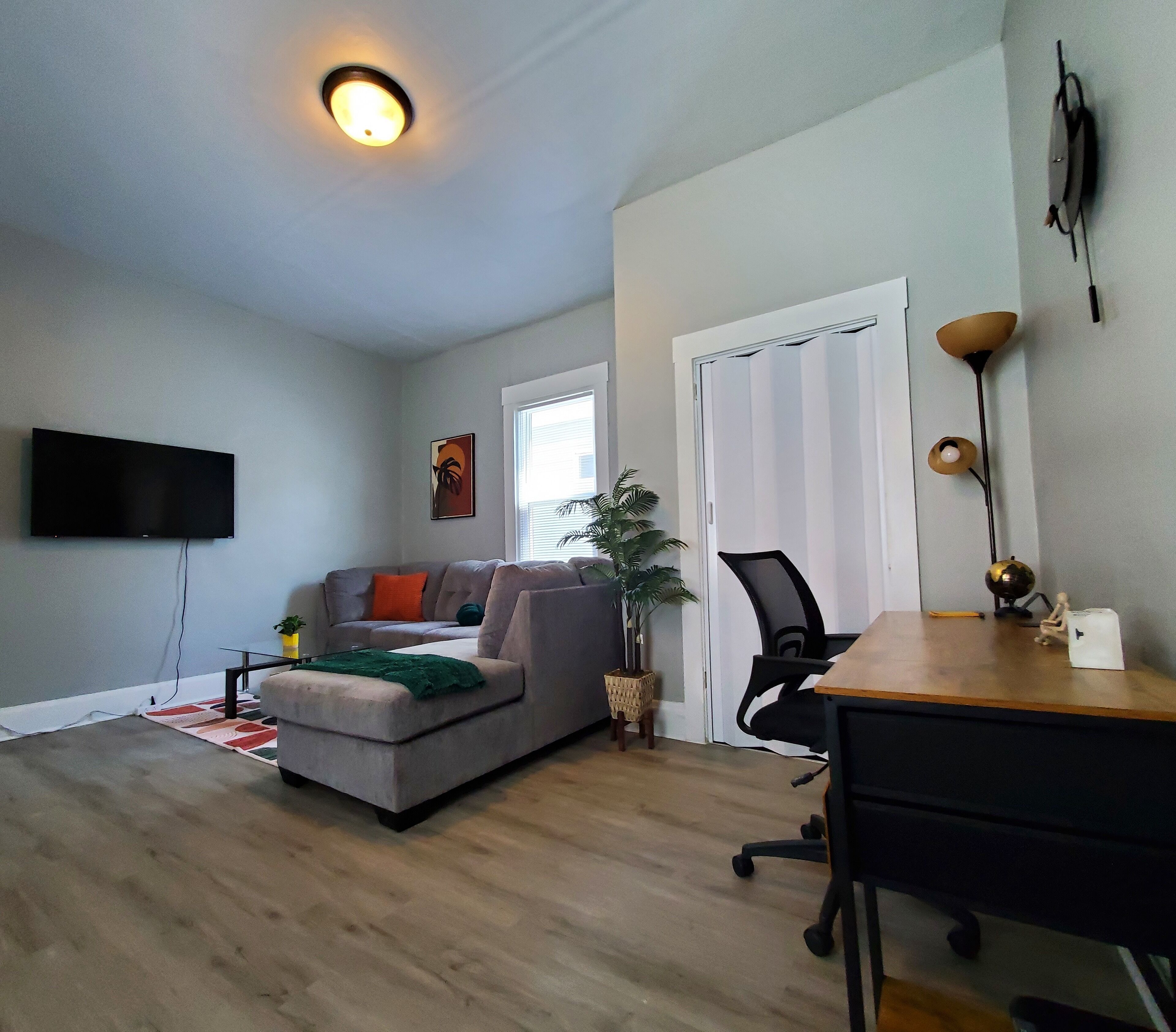 Un salon avec un canapé gris, un téléviseur à écran plat, un bureau en bois et une petite plante.