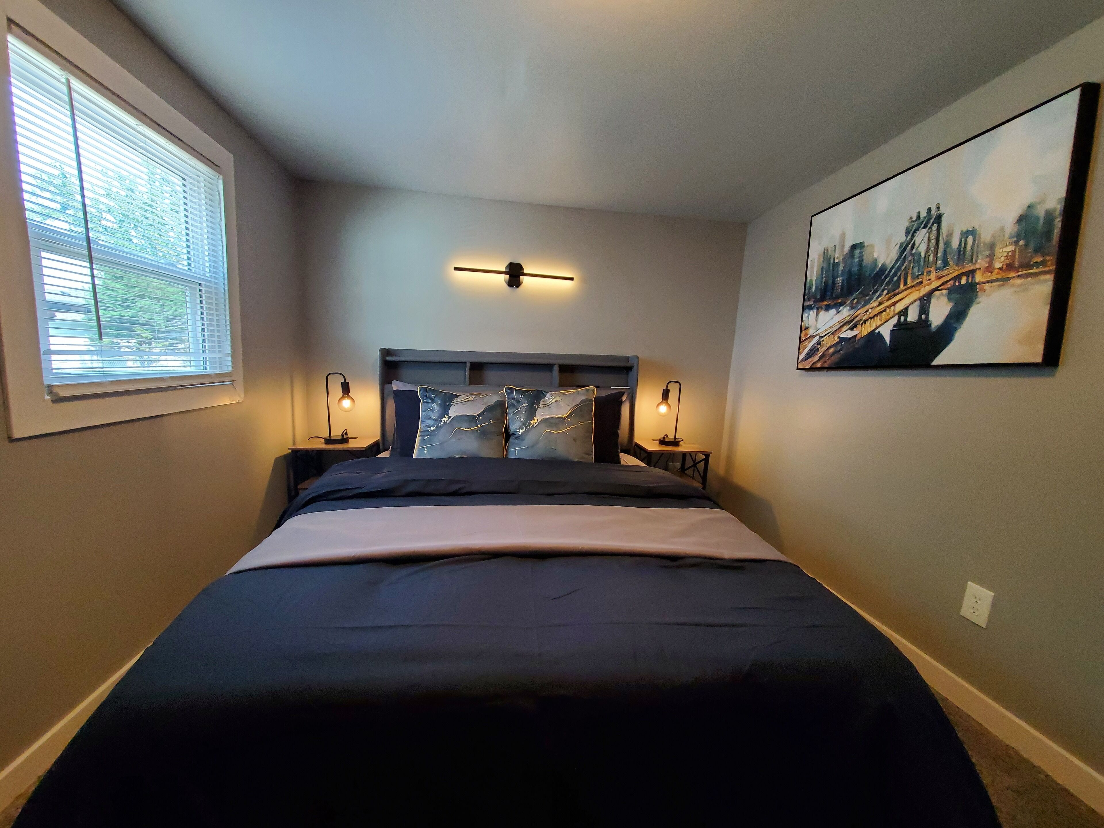 A neatly made bed with a headboard, two bedside lamps, and a painting on the wall.