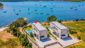 A modern two-story building with a swimming pool, surrounded by a lush landscape and a view of a calm sea with numerous boats.