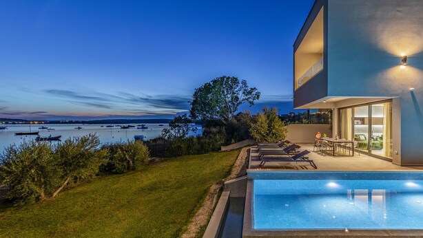 A modern house with a swimming pool, a patio with lounge chairs, and a view of the water and boats.
