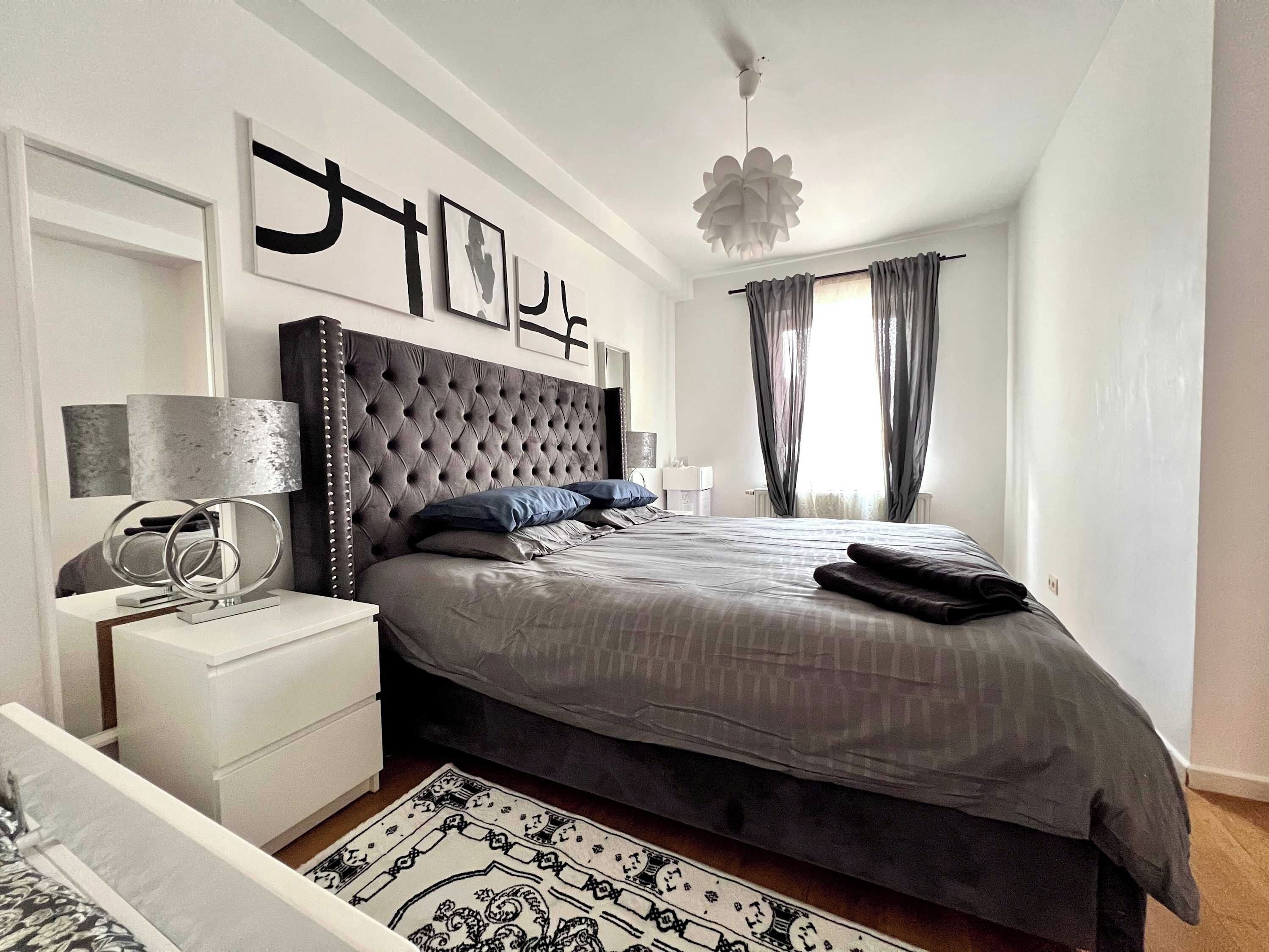 Een moderne slaapkamer met een gestoffeerd hoofdeinde, een groot bed, een nachtkastje met lamp en een decoratief vloerkleed.