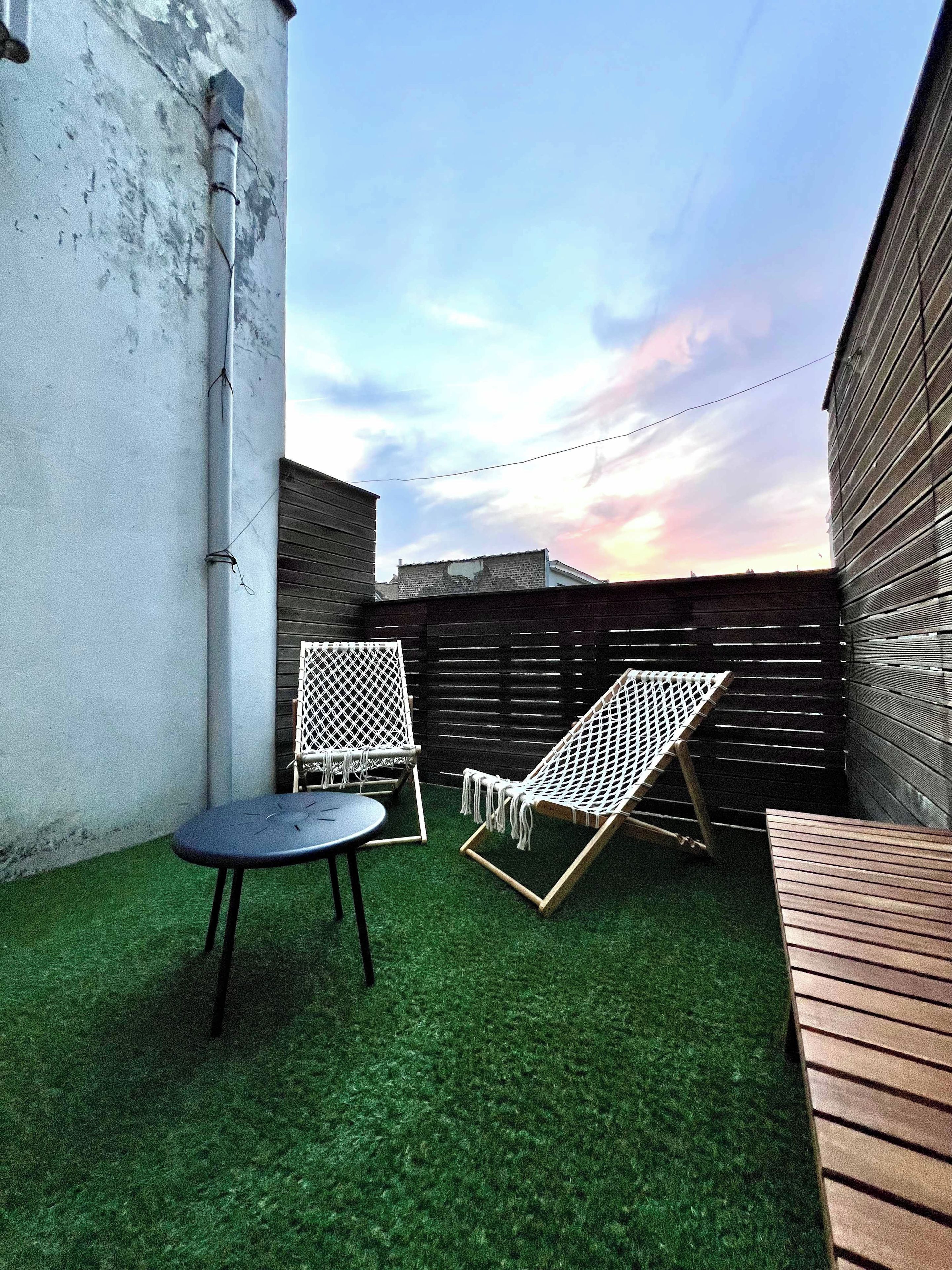 Een terras met twee witte stoelen en een tafel op kunstgras, omgeven door een houten hek en een gevel.