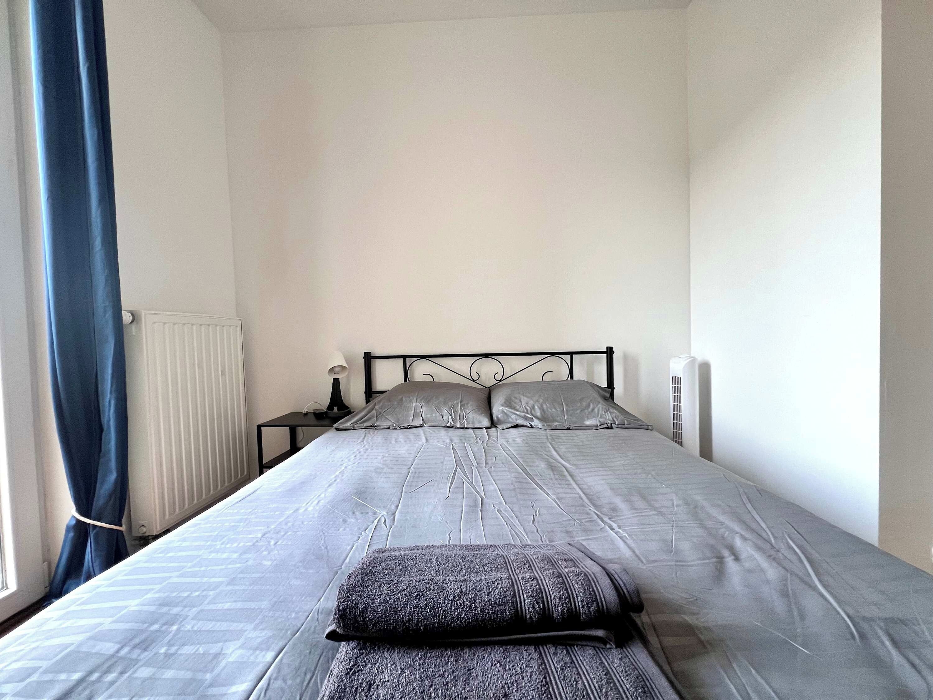 Een slaapkamer met een bed, nachtkastje, lamp, radiator en een raam met gordijnen.