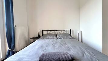 Een slaapkamer met een bed, nachtkastje, lamp, radiator en een raam met gordijnen.