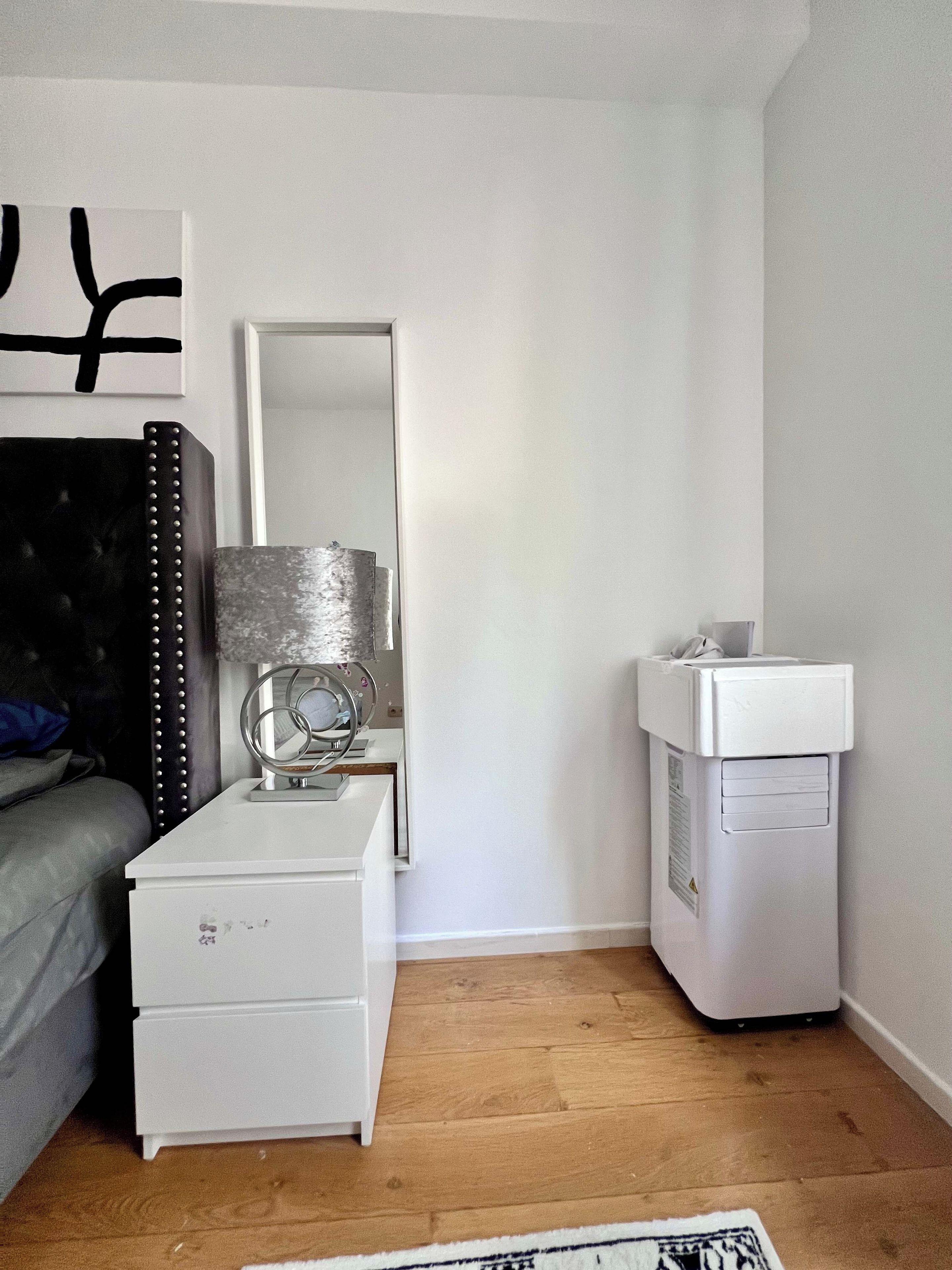 Een slaapkamer met een bed, een nachtkastje met een lamp, een spiegel, een dressoir en een witte prullenbak.