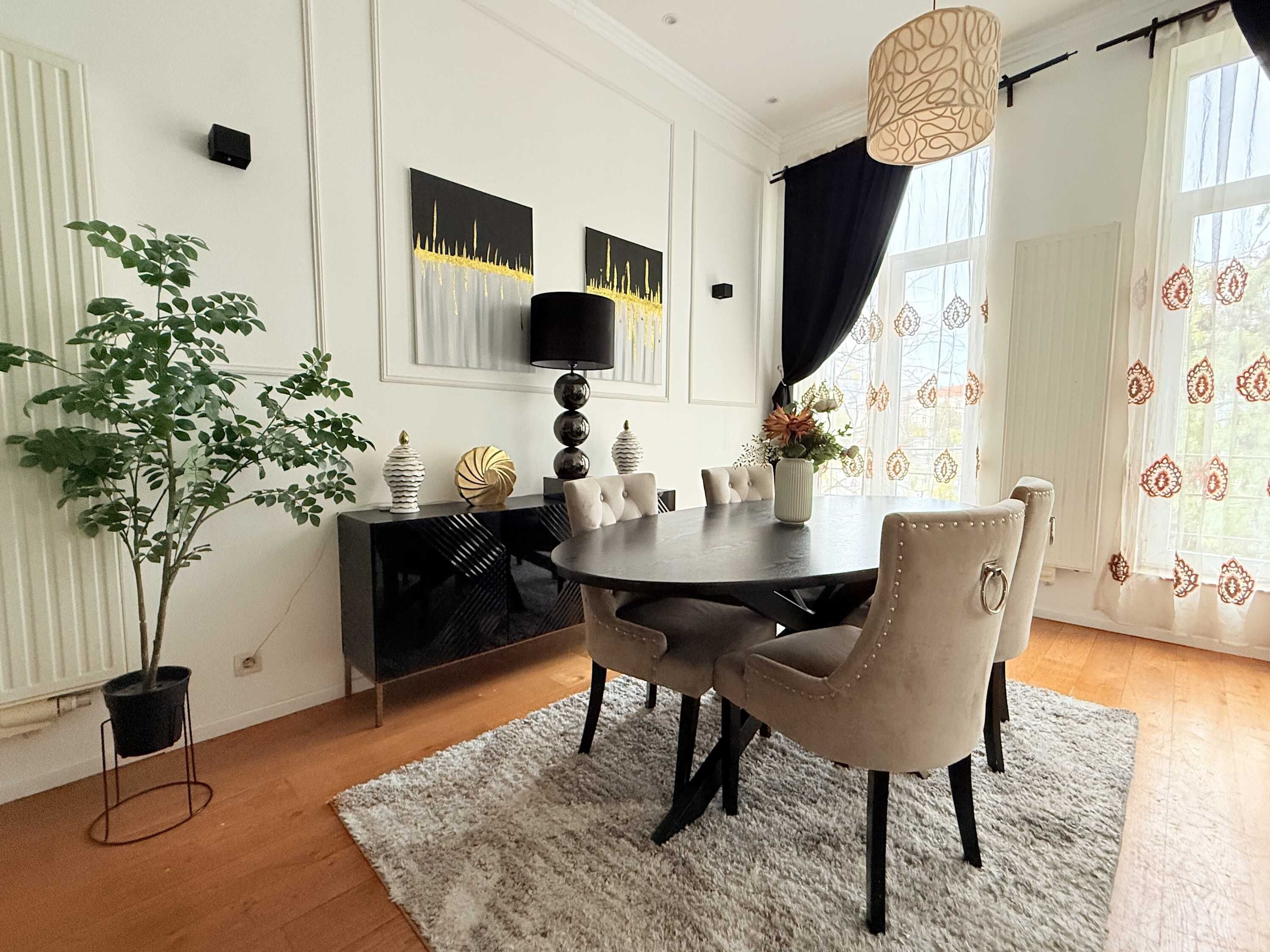 Een eetruimte met een ronde tafel, stoelen, een potplant, kunst aan de muur en een raam met gordijnen.