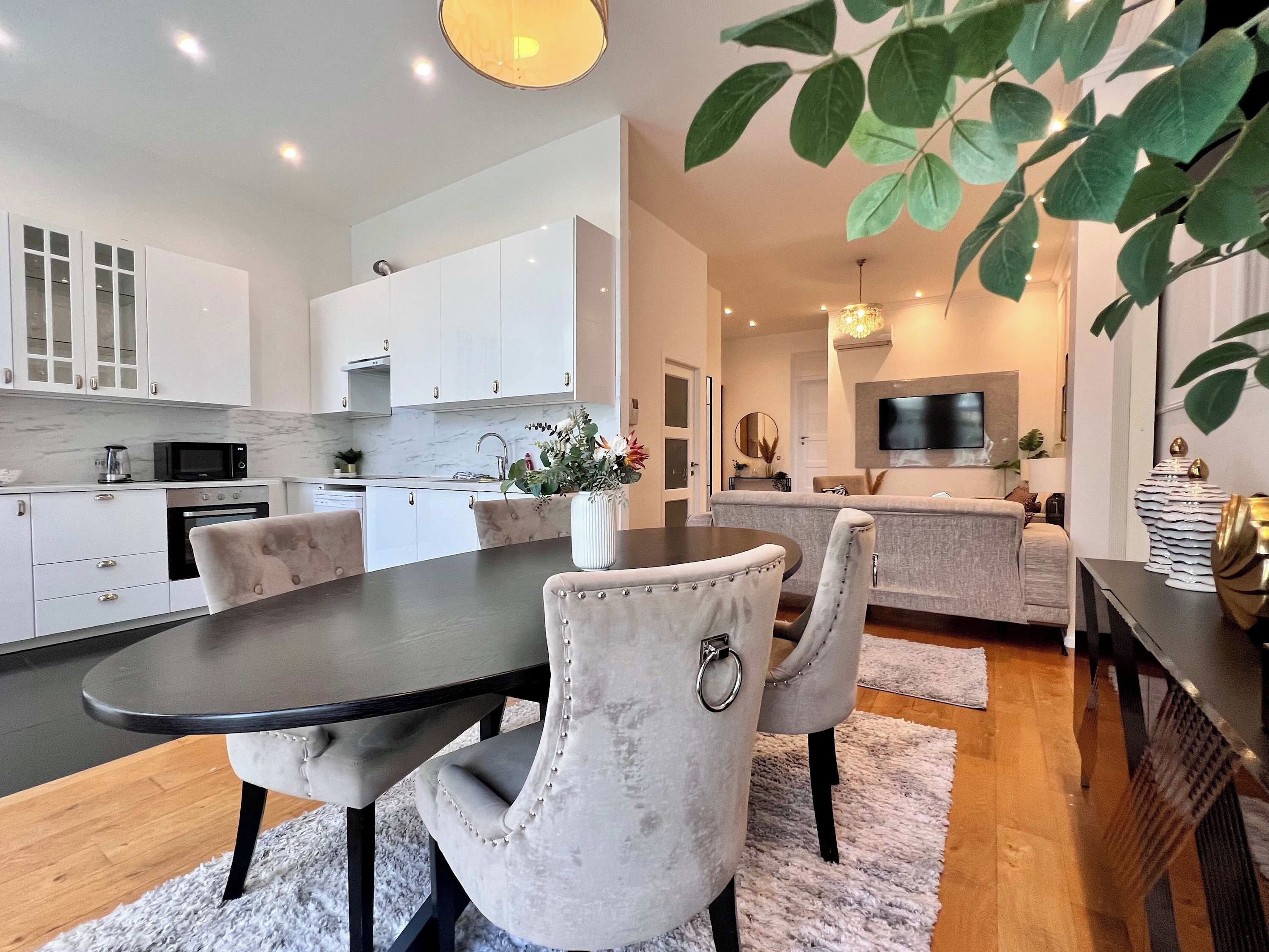 Een moderne keuken met witte kasten, een eethoek met een donkere tafel en gestoffeerde stoelen, en een zithoek met een bank en een flatscreen-televisie.