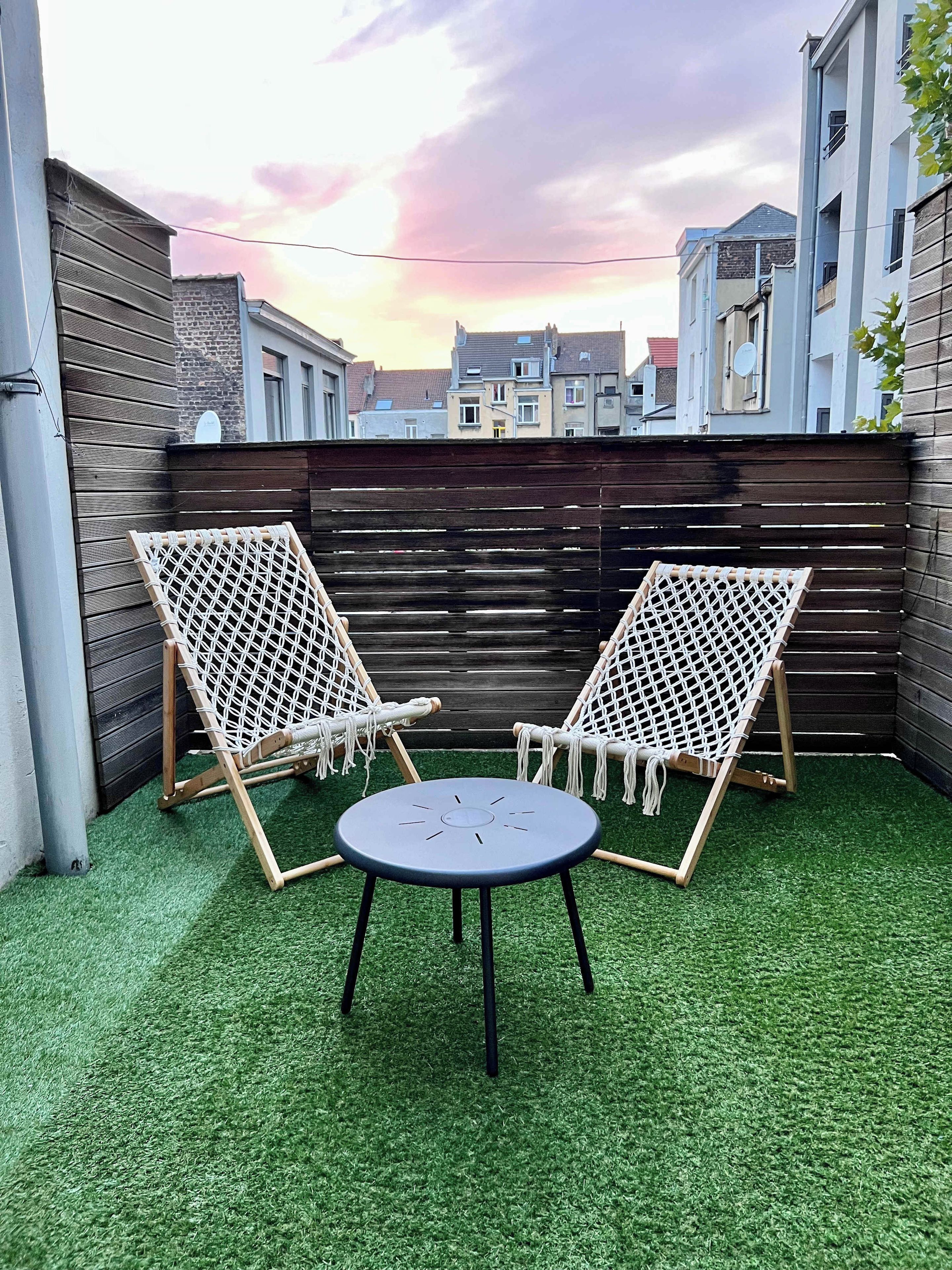 Een terras met twee witte rieten stoelen en een klein rond tafeltje op kunstgras, omgeven door een houten hek.