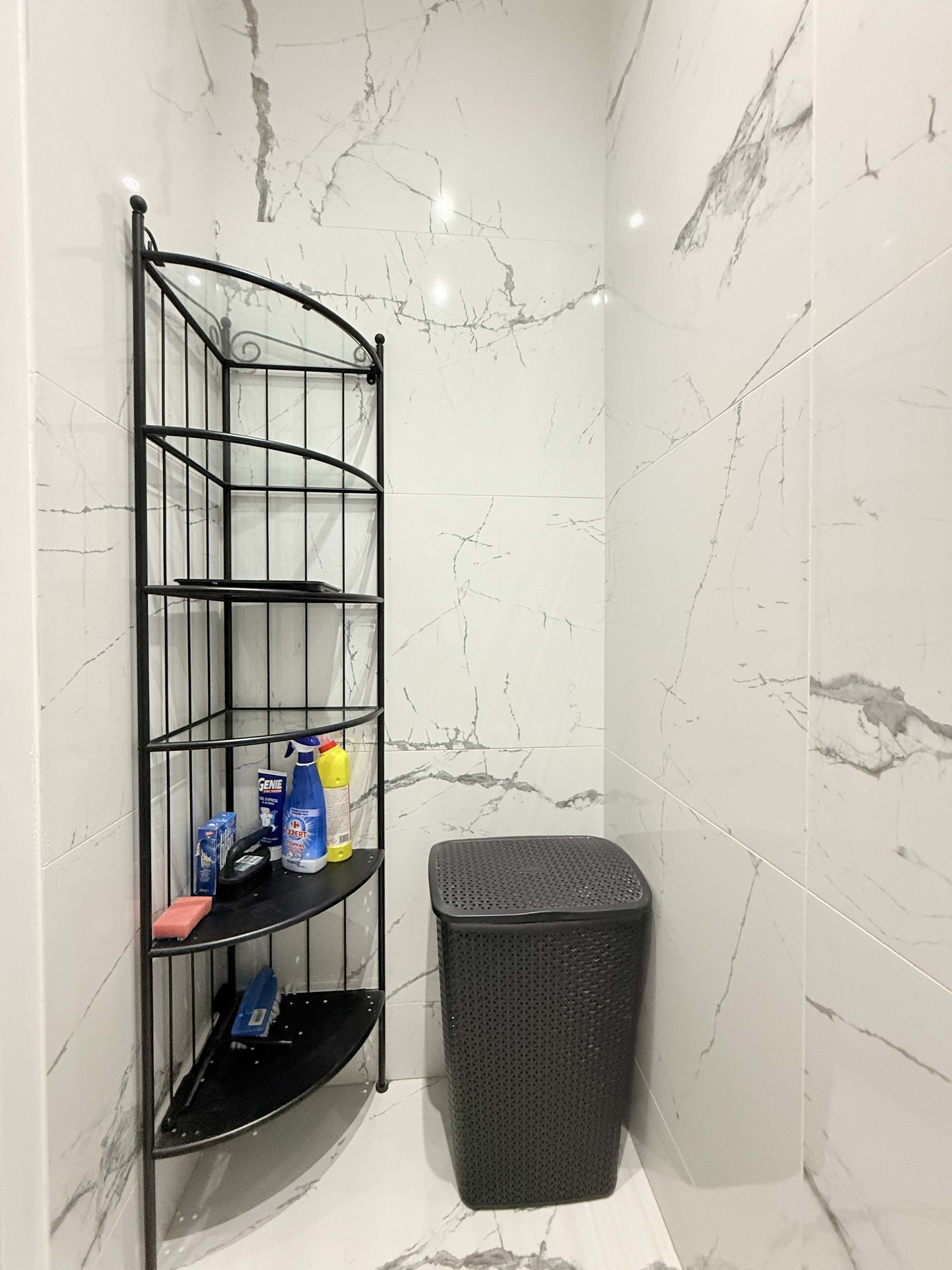 Een badkamer met een hoekplankje met toiletartikelen, een zwarte prullenbak en wanden met een marmerprint.