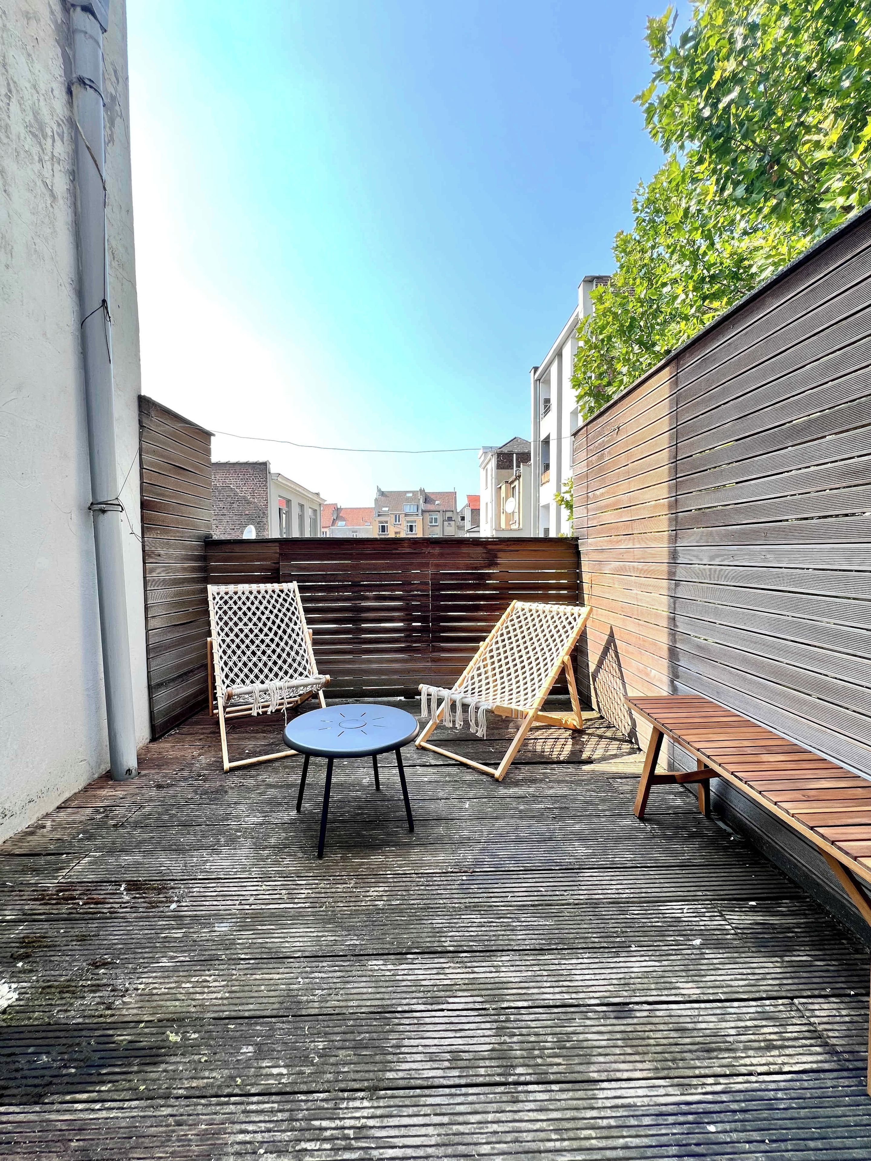 Un espace extérieur aménagé avec une table ronde et deux chaises, un banc en bois et une clôture en métal.