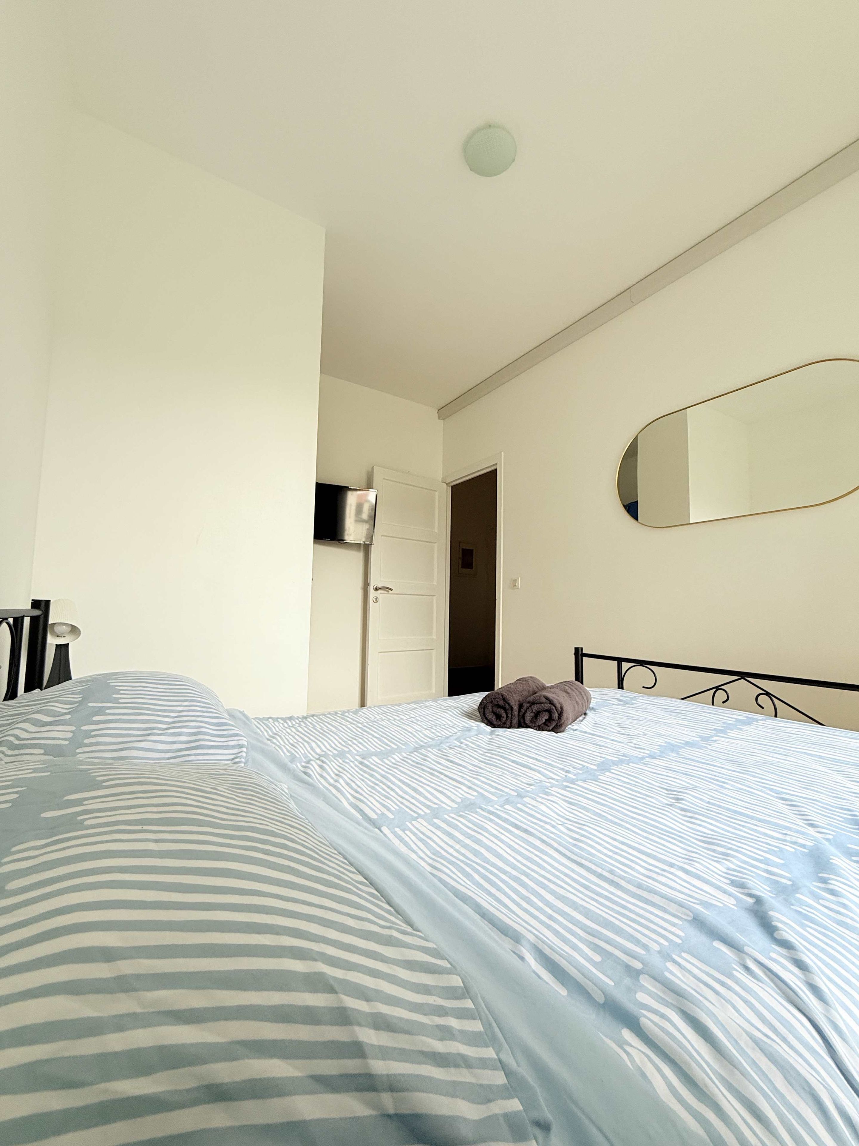 A bedroom with a bed, a bedside table, a mirror, and a door leading to another room.