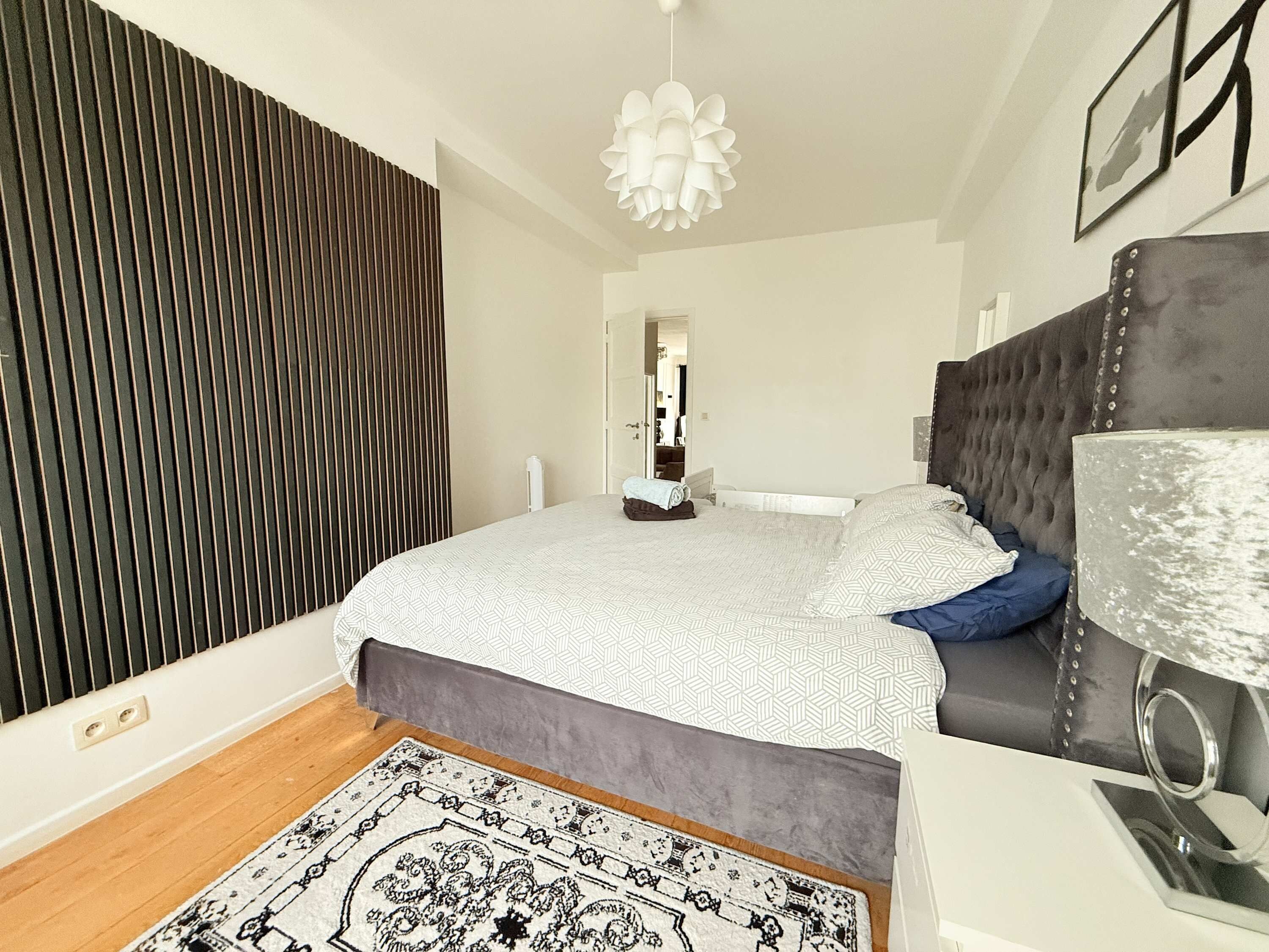 Een slaapkamer met een bed, een bedlamp, een kamerkleed met een patroon en een donker gestoffeerd hoofdeinde.
