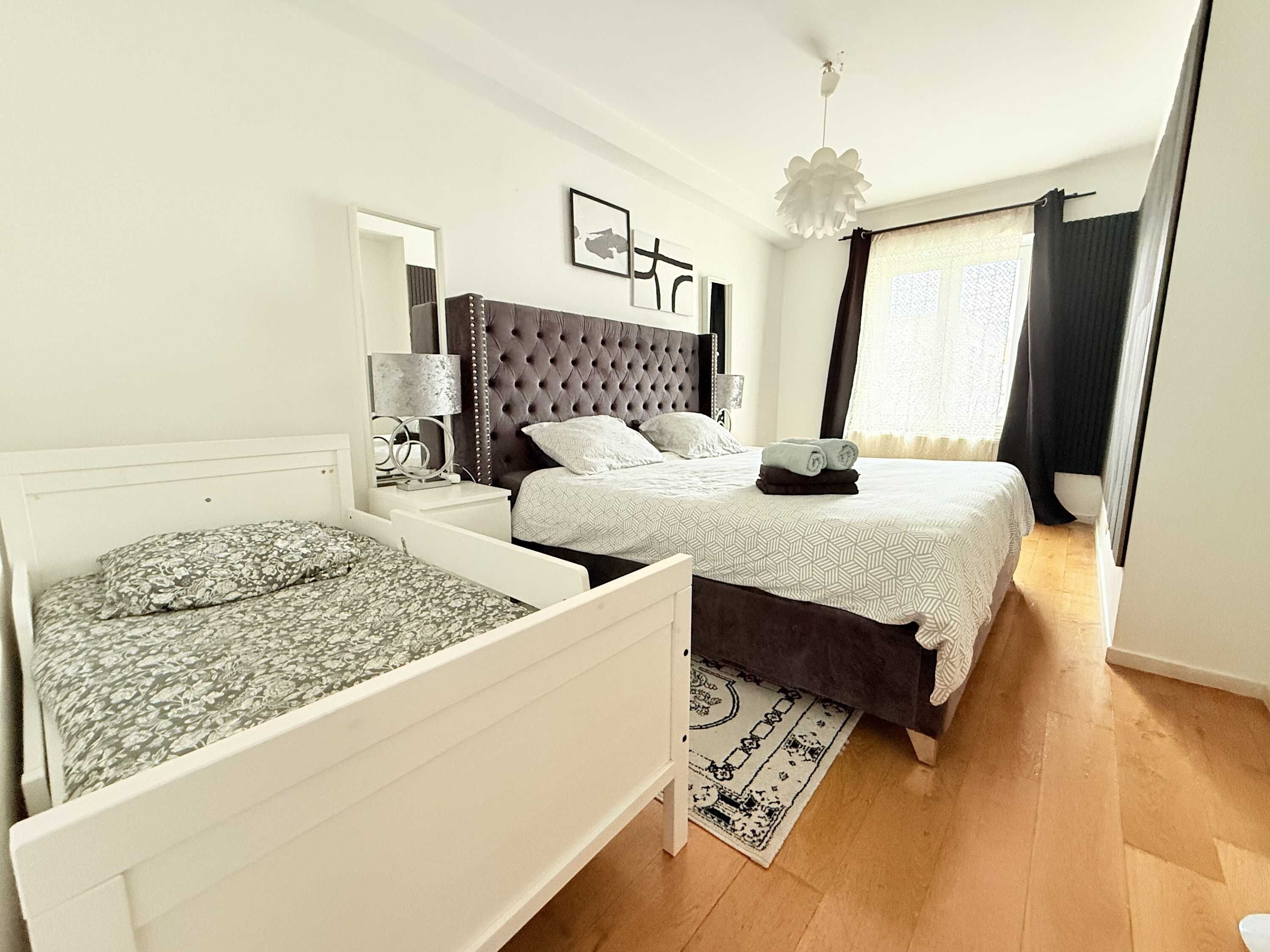 A bedroom with a large bed, a smaller bed, a nightstand, a lamp, a mirror, and a window with curtains.