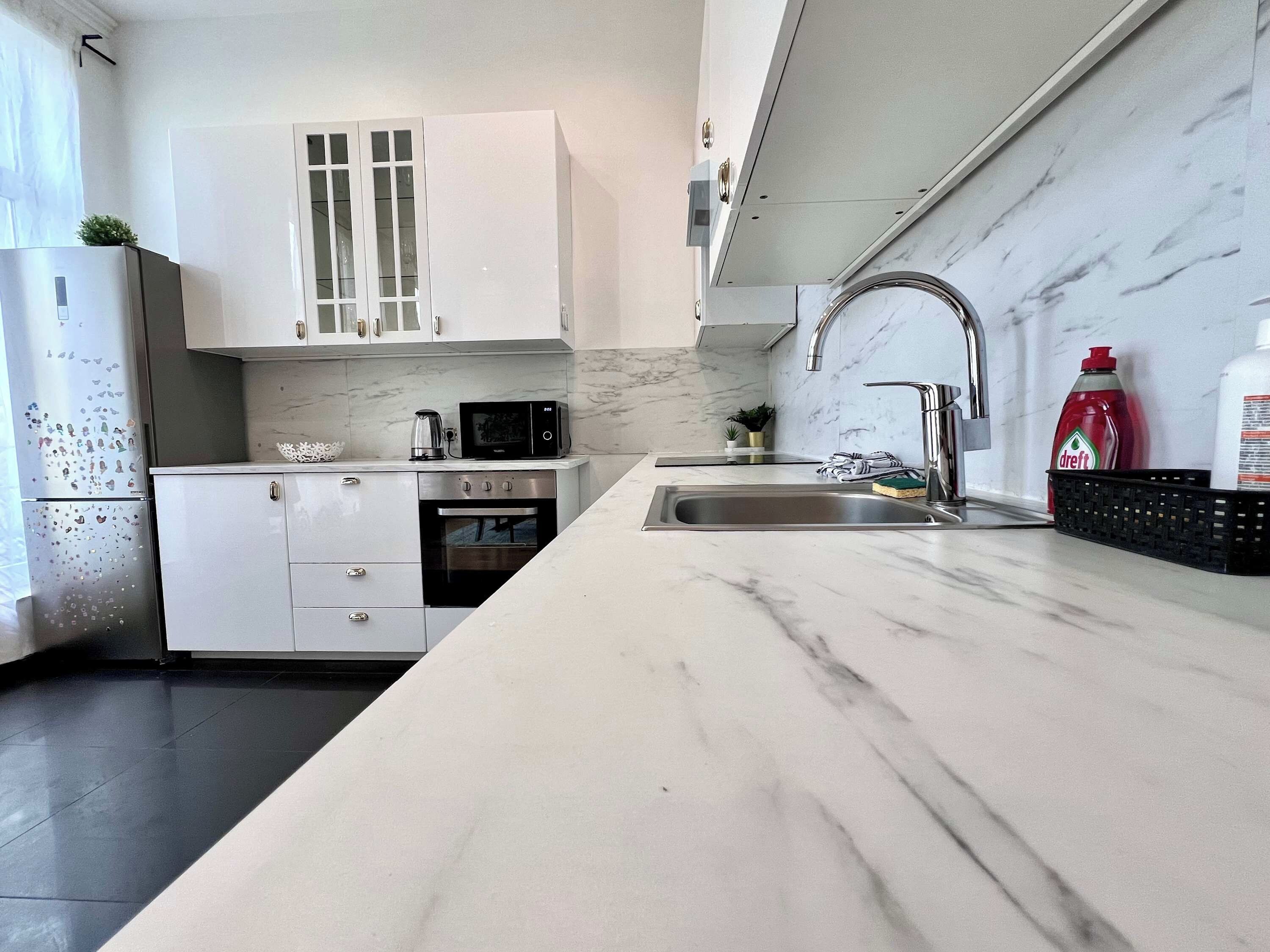 Een moderne keuken met witte kasten, een roestvrijstalen koelkast, een magnetron en een wastafel met een marmeren aanrecht.
