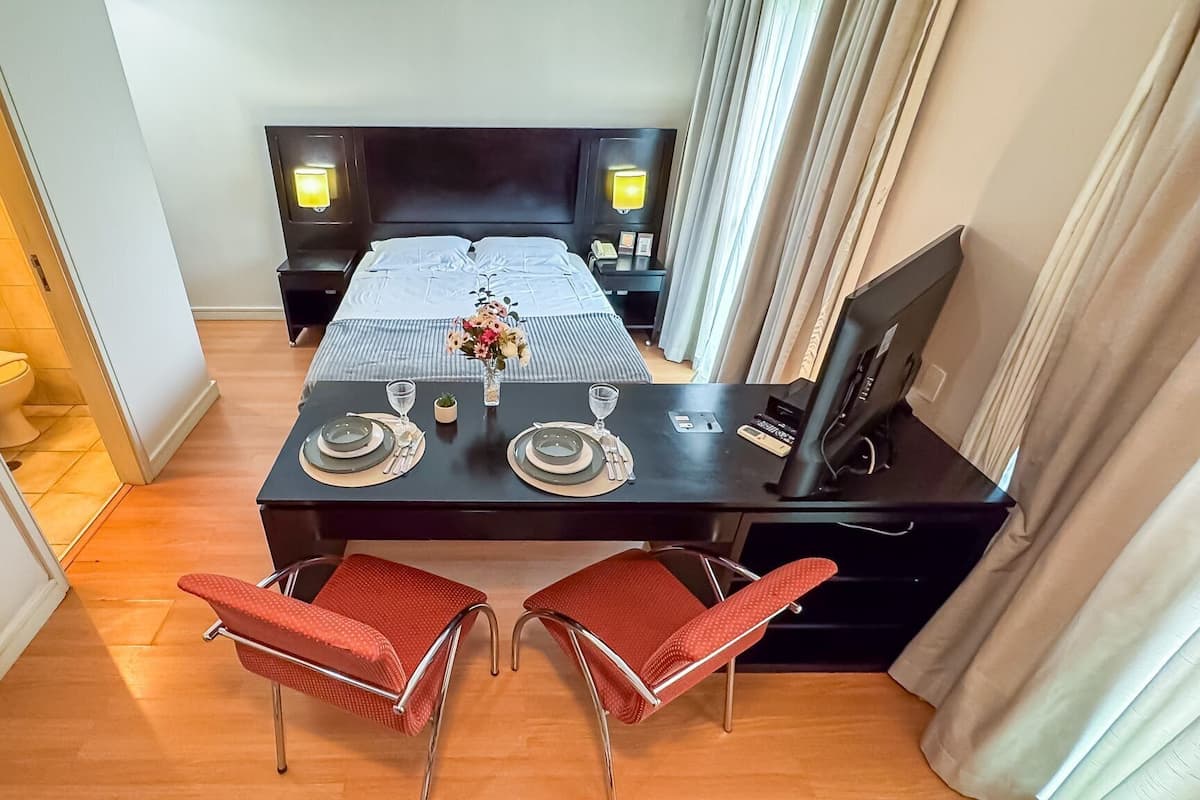 Une chambre d’hôtel avec un lit, une petite table mise pour deux personnes avec des assiettes, des verres et un vase de fleurs. Il y a deux chaises rouges et un téléviseur posé sur un meuble.