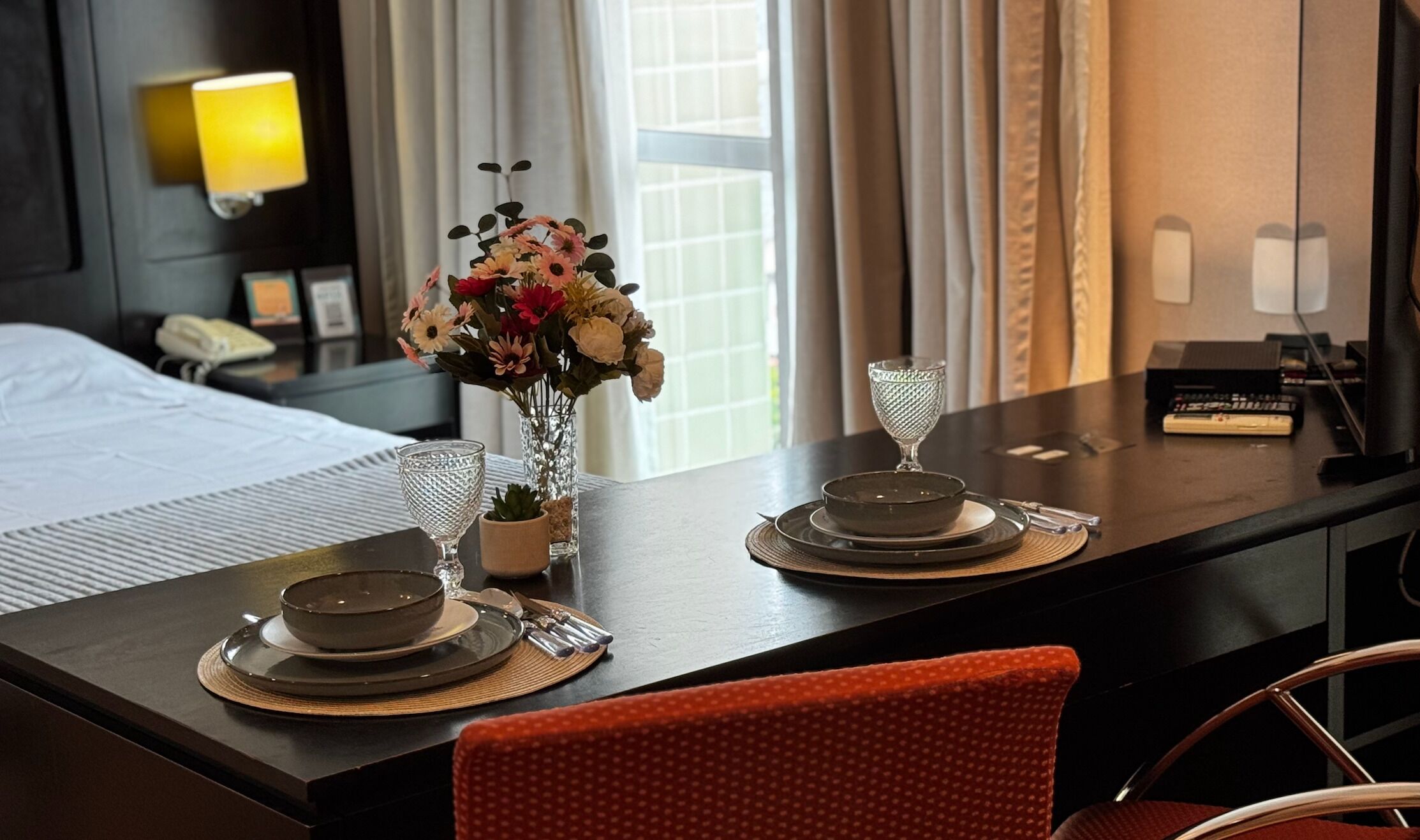 Ett hotellrum med ett välordnat matbord dukat för två personer, en vas med blommor, en sänglampa och en säng med ett vitt överkast.
