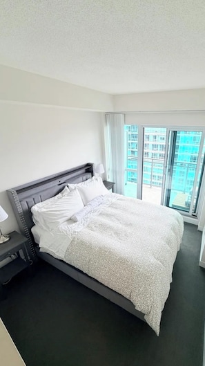 Hotellihuone, jossa on suuri sänky, yöpöytä, lamppu ja ikkunasta avautuva näkymä rakennukseen.