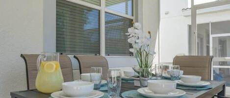 A dining table set with plates, glasses, and a pitcher of lemonade, with chairs and a window in the background.