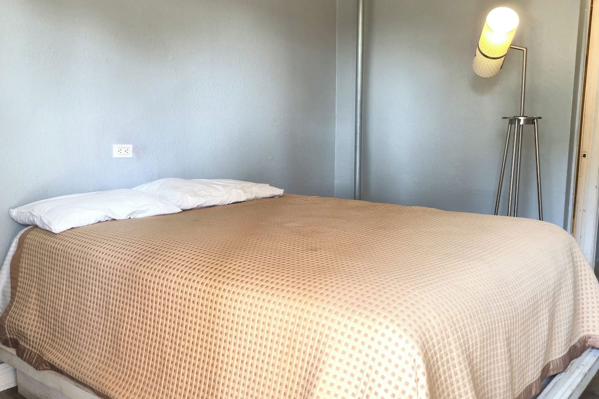 Un lit simple avec une couvre-lit beige à motifs, un oreiller blanc et une lampe de sol dotée d’un abat-jour cylindrique.