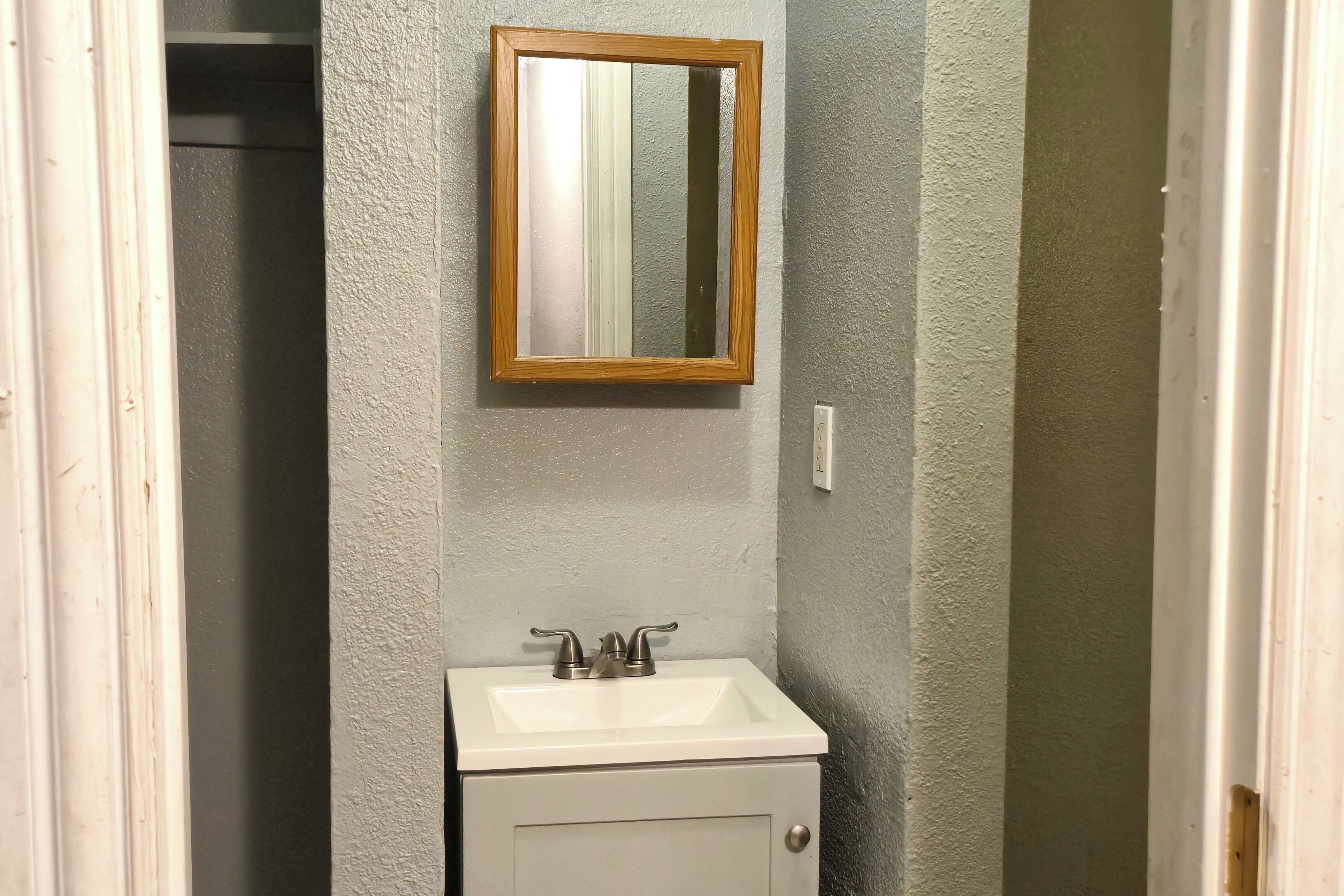 Un baño con un lavamanos blanco, un espejo con marco de madera y una pared gris con textura.