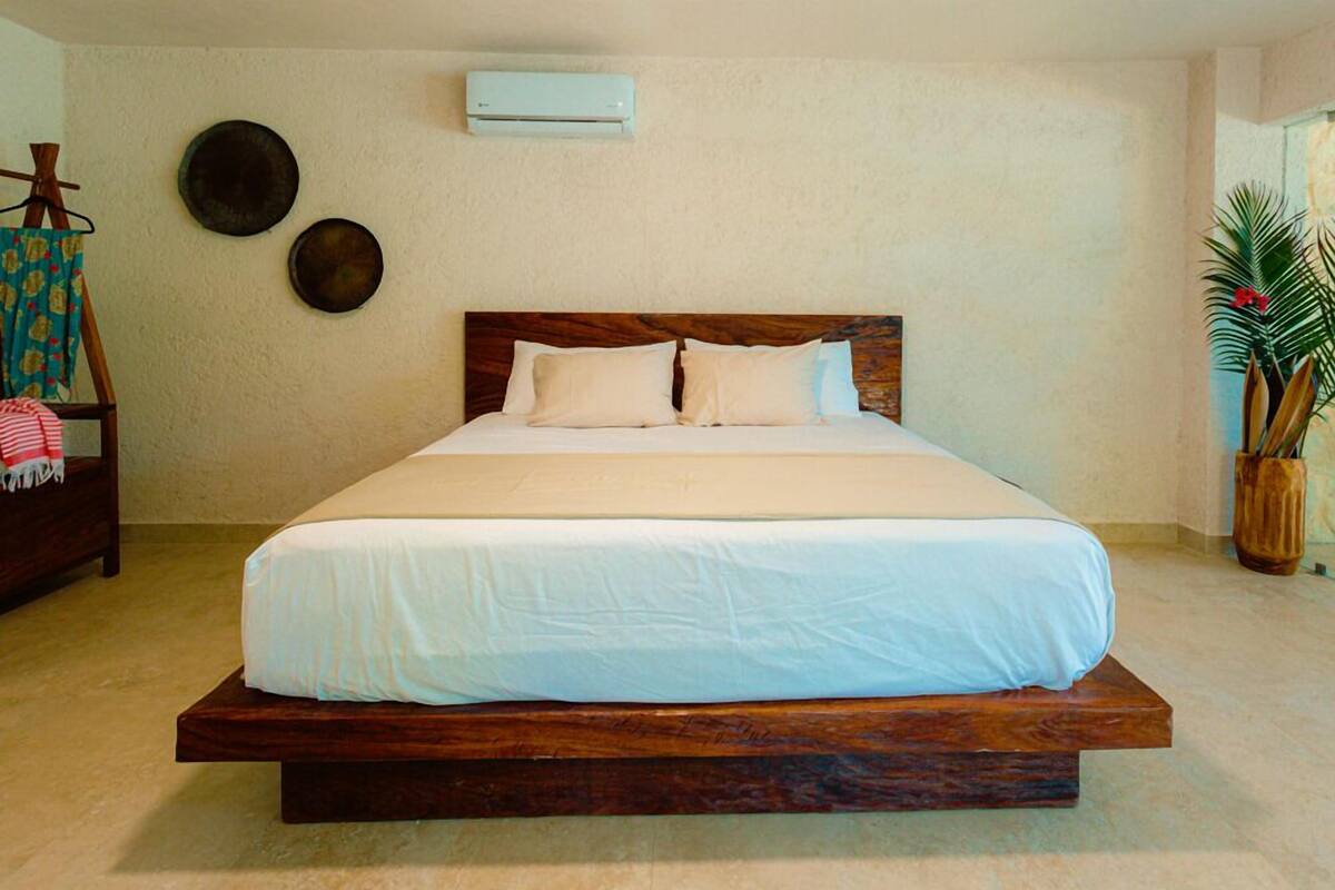 Ein großes Bett mit Holzrahmen und weißer Bettwäsche, eine wandmontierte Klimaanlage und ein Blumentopf auf der rechten Seite.