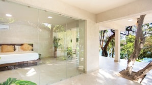 A modern hotel room with a glass partition, a bed with pillows, a potted plant, and a view of trees and sky. - Corrales Bay Resort (Los Corrales)