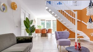 Una sala moderna con un sofá gris, un sillón púrpura y una zona de comedor con sillas naranjas. Una escalera blanca con barandilla de vidrio conduce a un nivel superior.