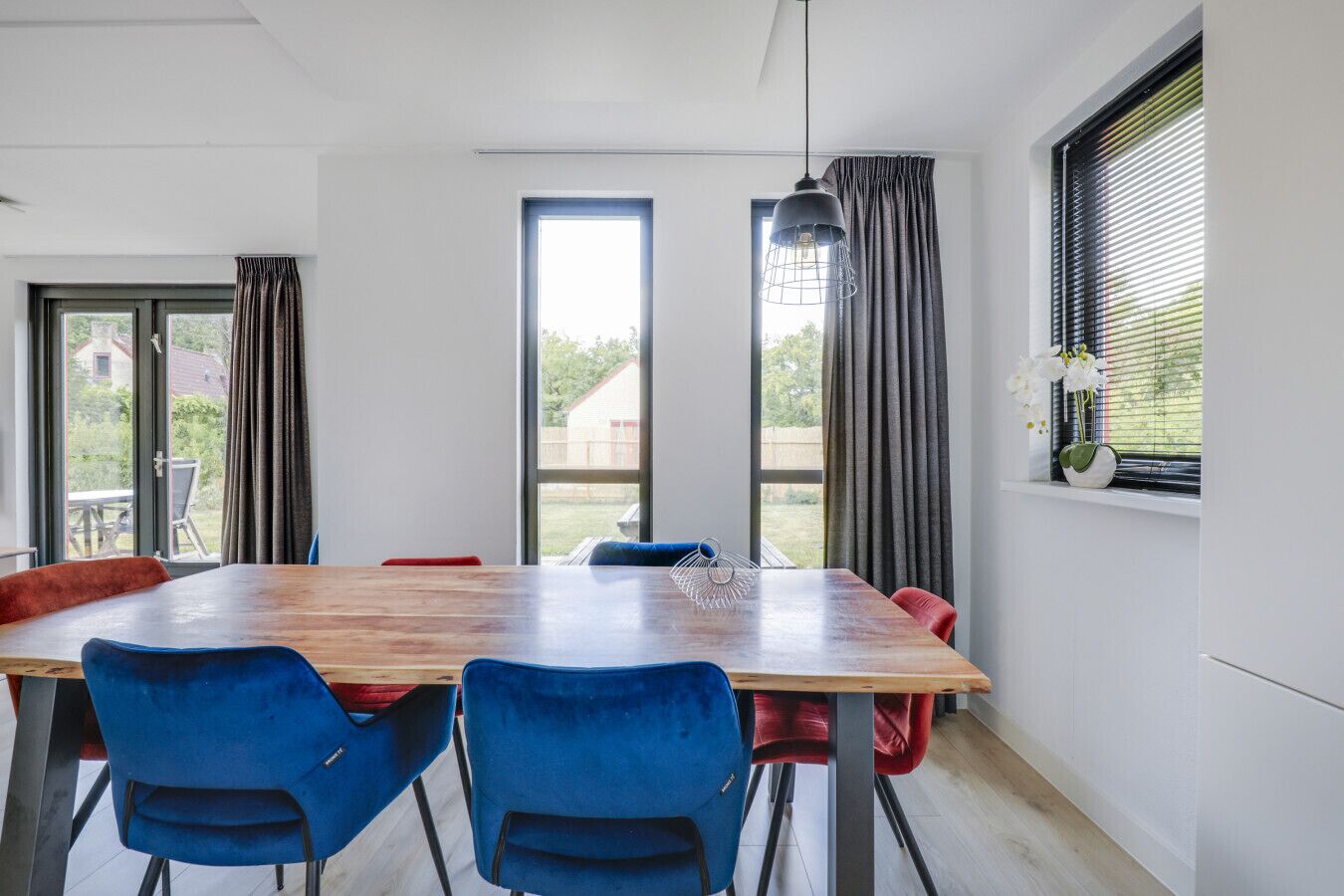 A dining area with a wooden table and blue chairs, large windows with curtains, and a hanging light fixture.
