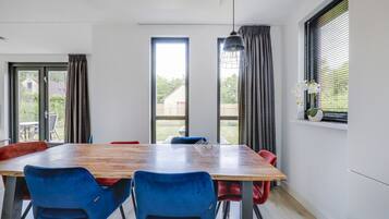A dining area with a wooden table and blue chairs, large windows with curtains, and a hanging light fixture.