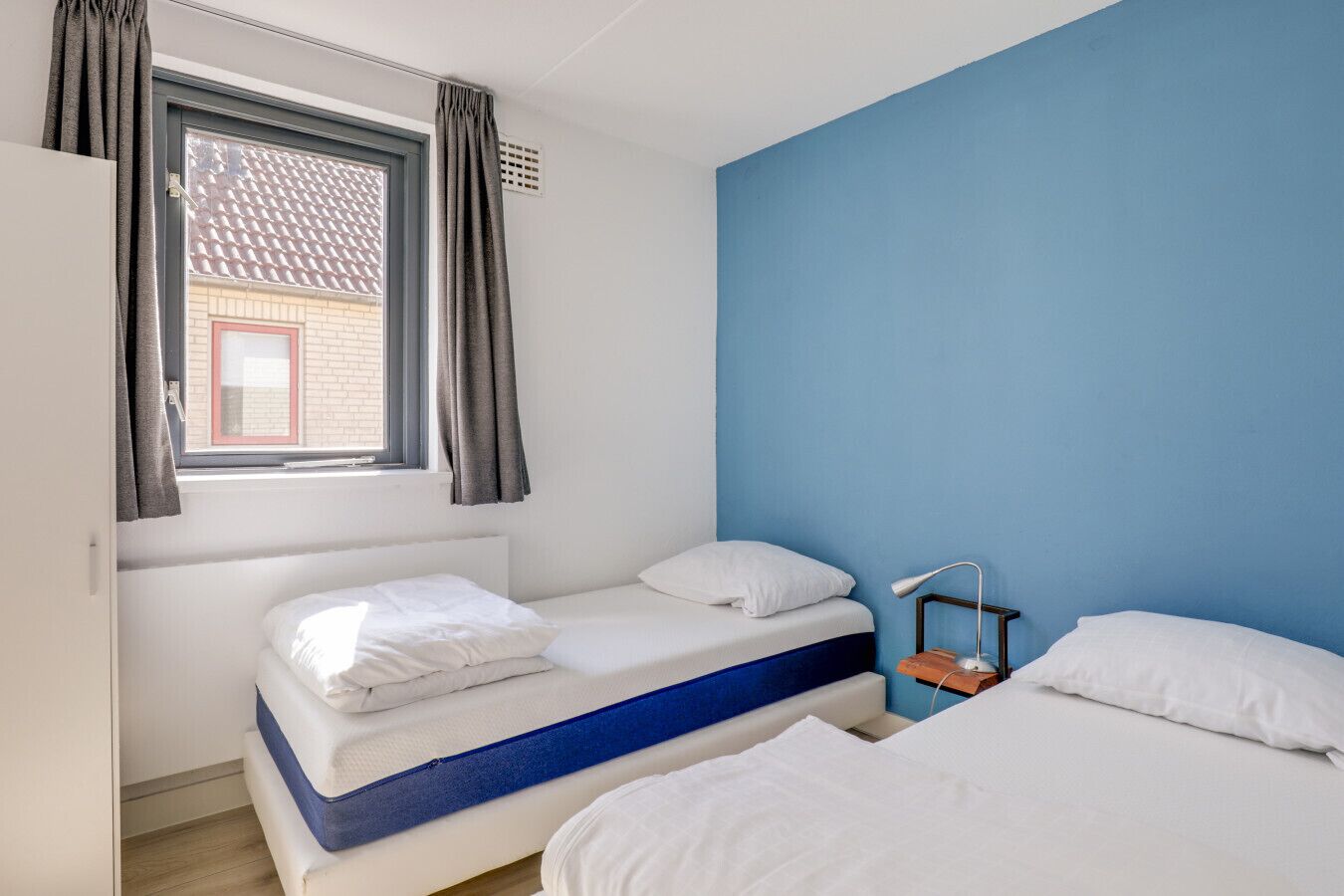 A room with two beds, a window with curtains, a desk with a lamp, and a blue wall.