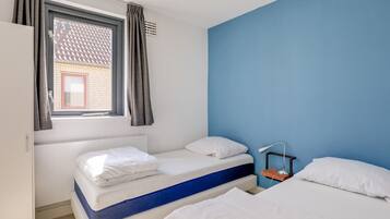 A room with two beds, a window with curtains, a desk with a lamp, and a blue wall.
