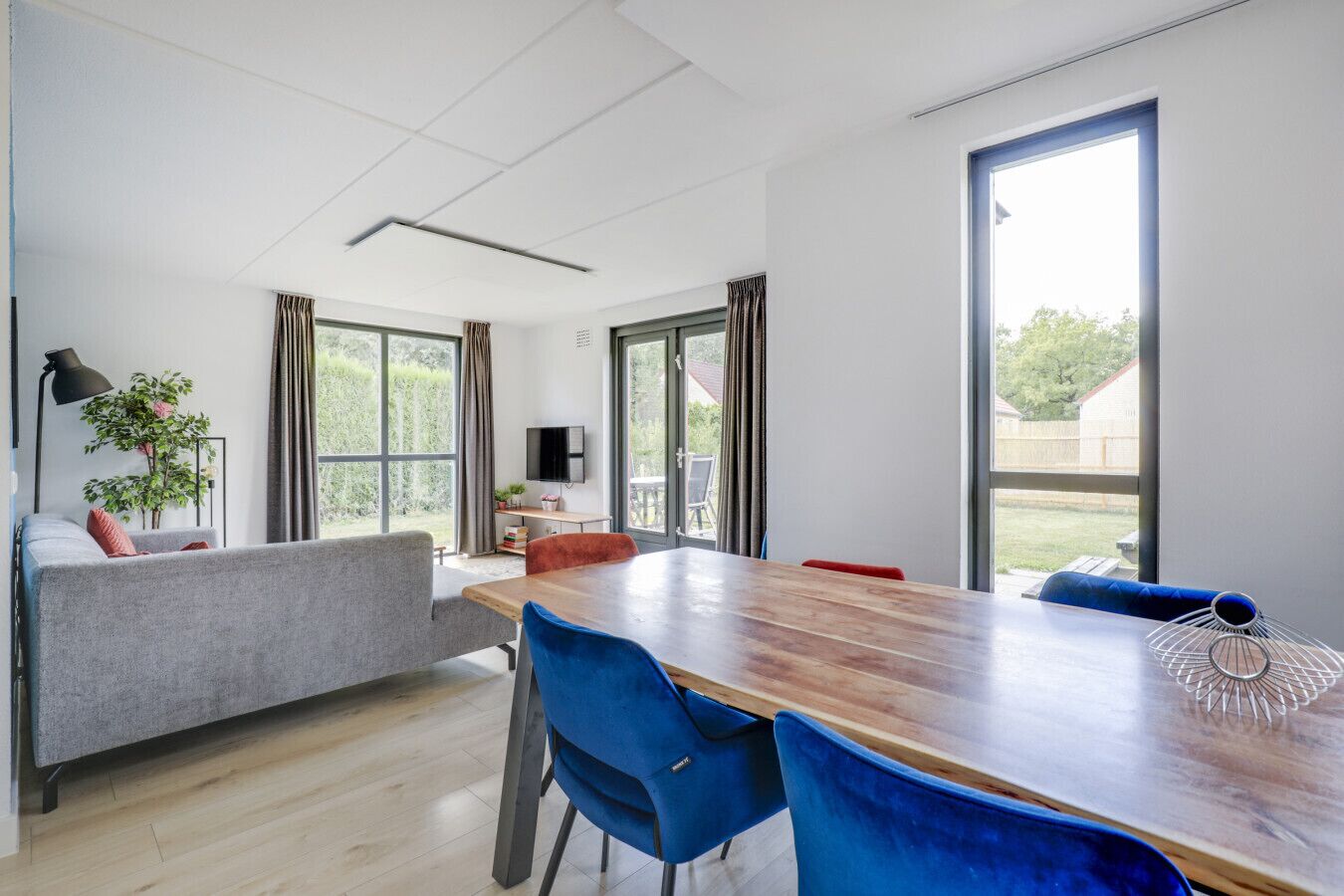 Een moderne woonkamer met een houten eettafel, blauwe stoelen, een grijze bank en grote ramen met uitzicht op de omgeving.