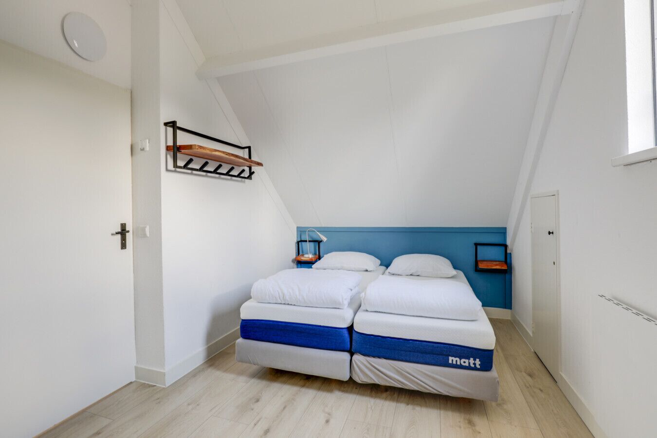 Een kleine, moderne slaapkamer met een schuine plafond, twee bedden met wit en blauw beddengoed, een wandplank en een raam.