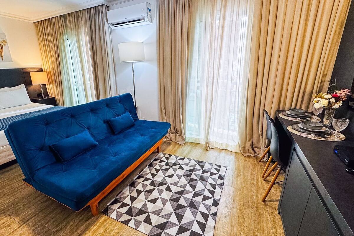 A hotel room with a blue sofa, a bed with white linens, a geometric patterned rug, and a dining area with a table set for two.